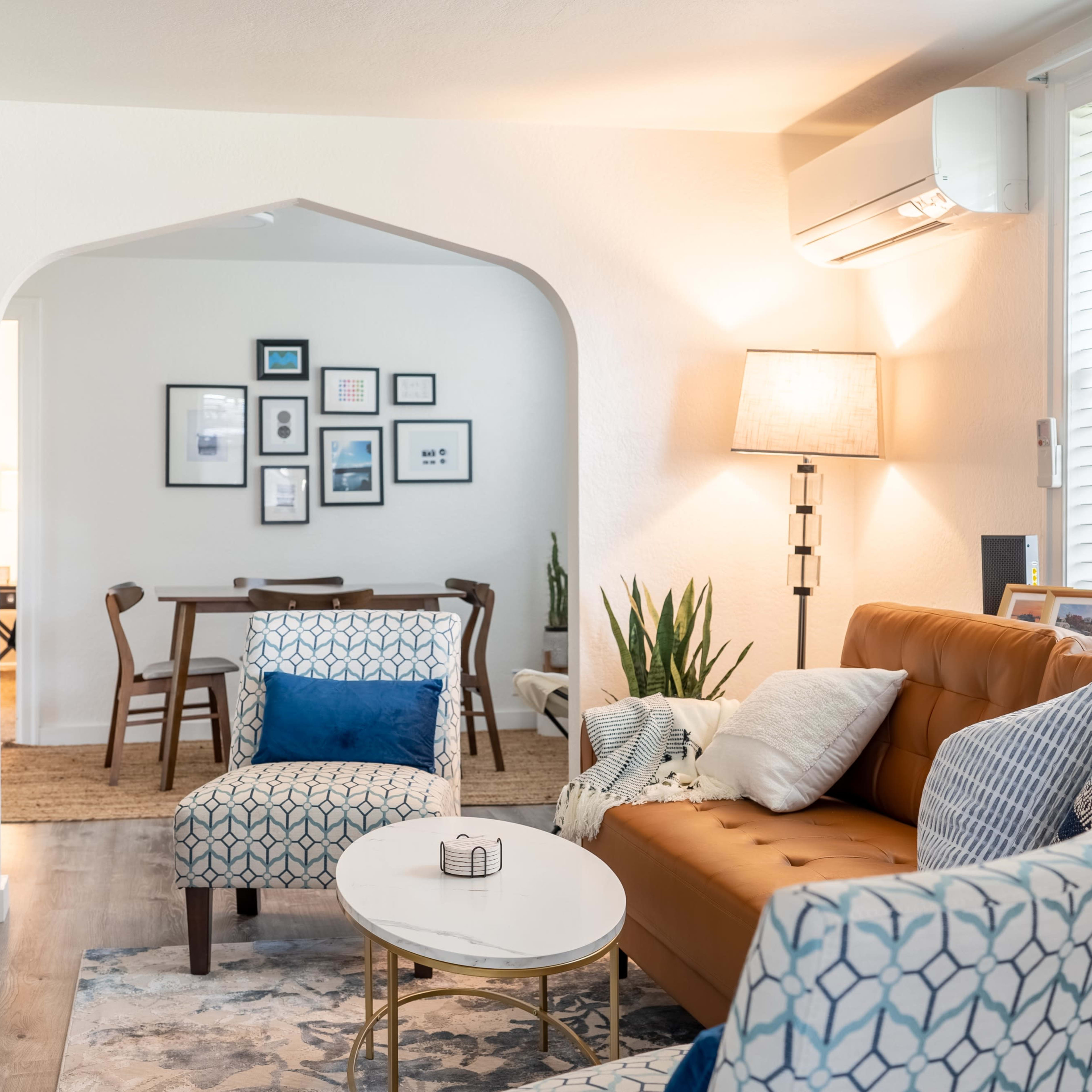 A living room with a brown leather couch, two patterned chairs, a round coffee table, and an archway leading to a dining area.