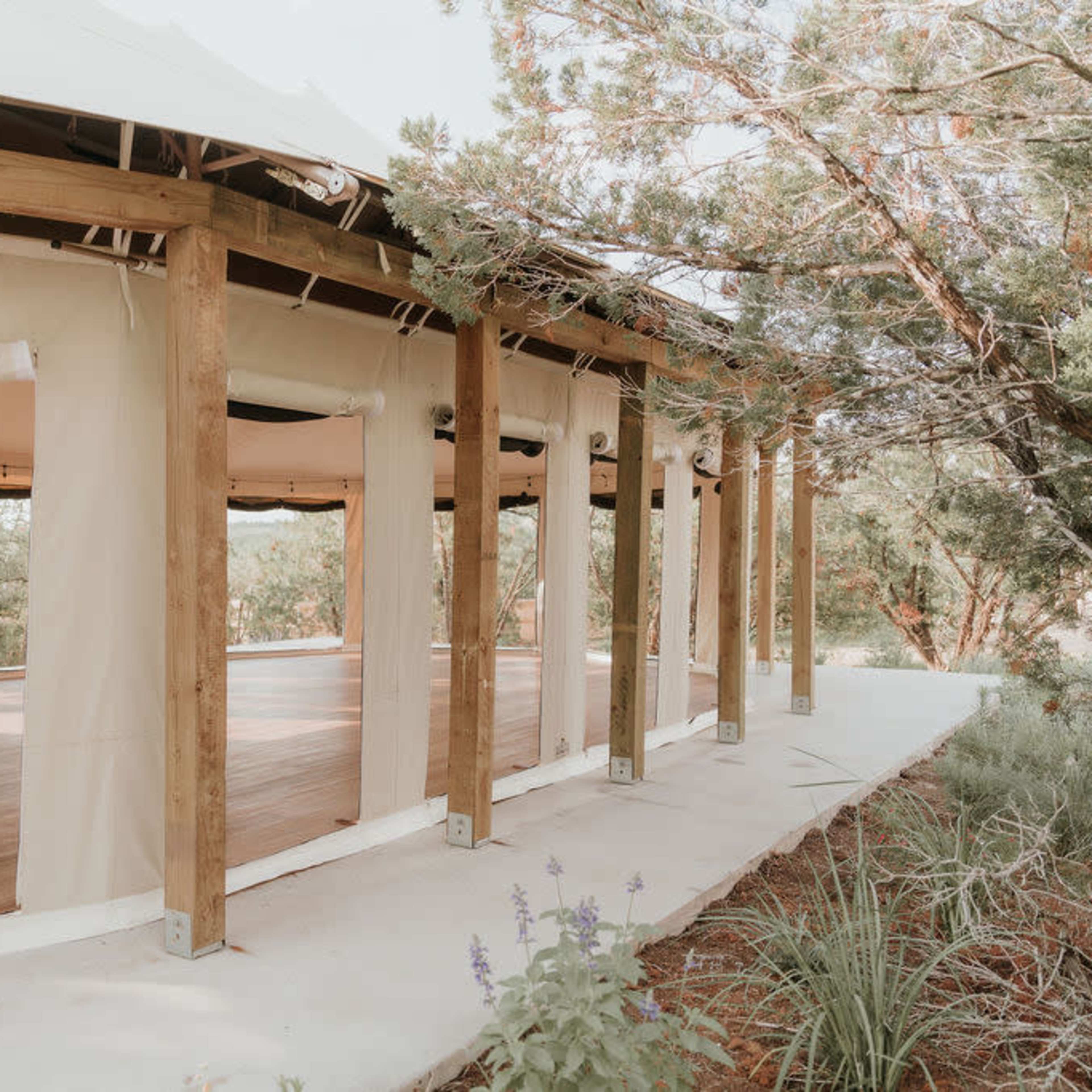 A modern, partially open structure with wooden beams and large fabric walls sits among greenery, featuring a spacious wooden deck and interior cabinetry.