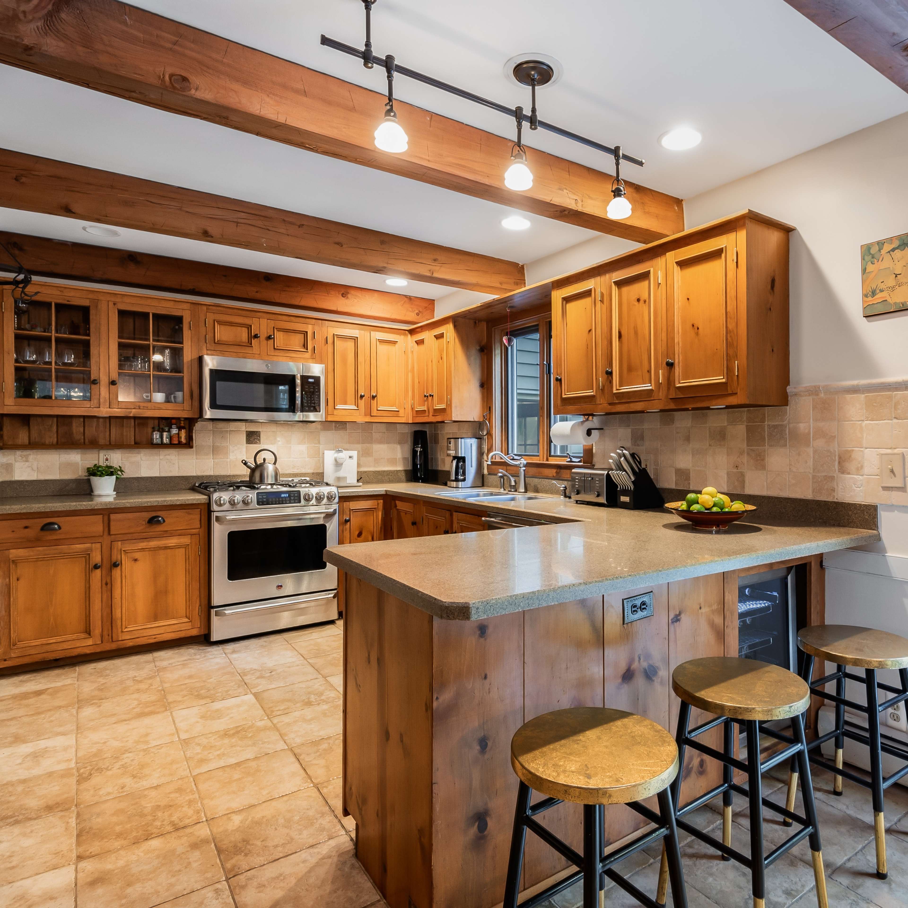 A kitchen with wooden cabinets and a granite island, equipped with stainless steel appliances and three metal stools.