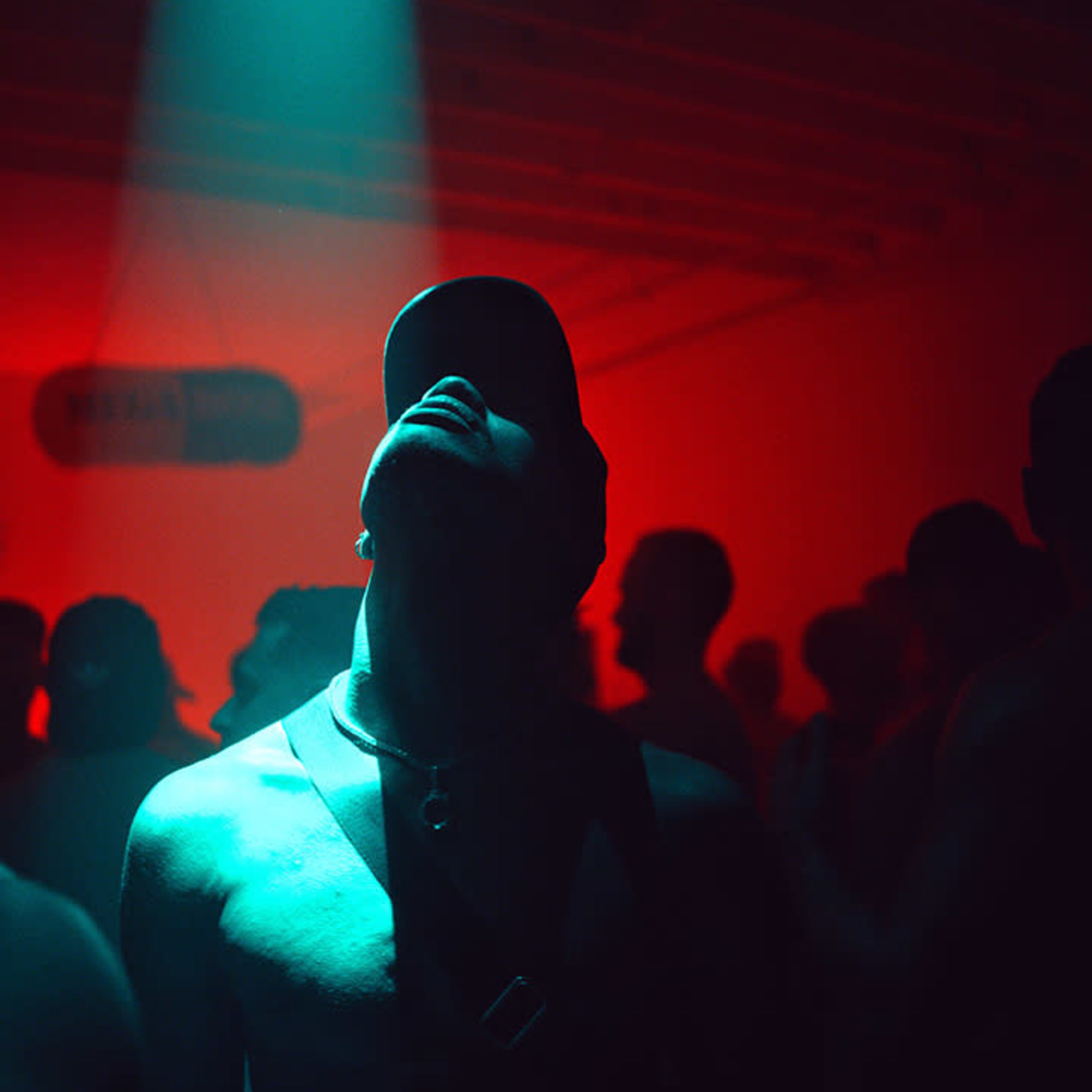 A man gazes upward in a dimly lit room illuminated by a blue spotlight, surrounded by other partygoers.