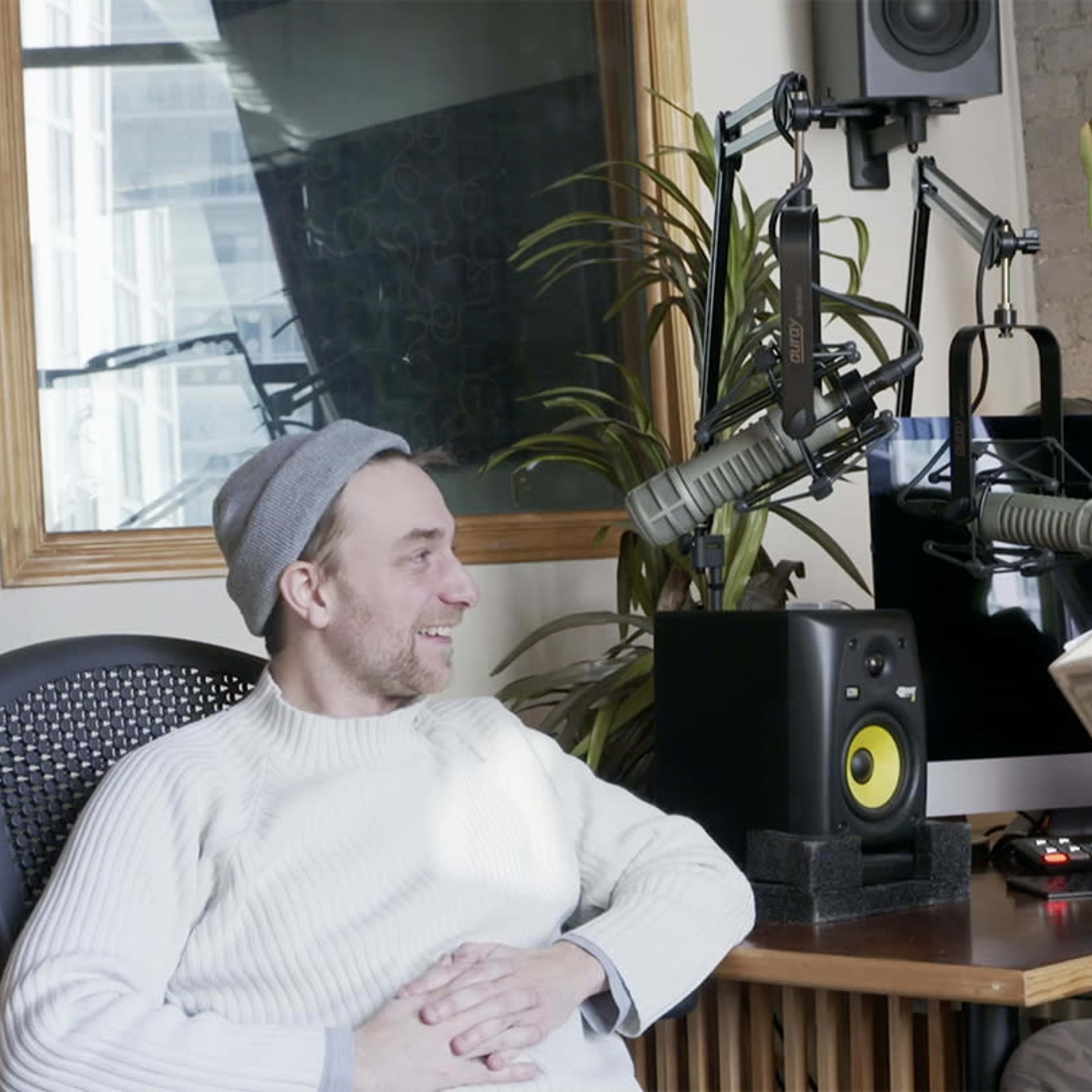 Two men sit in a studio setting, one reading from a book while the other listens, surrounded by audio equipment and a plant.