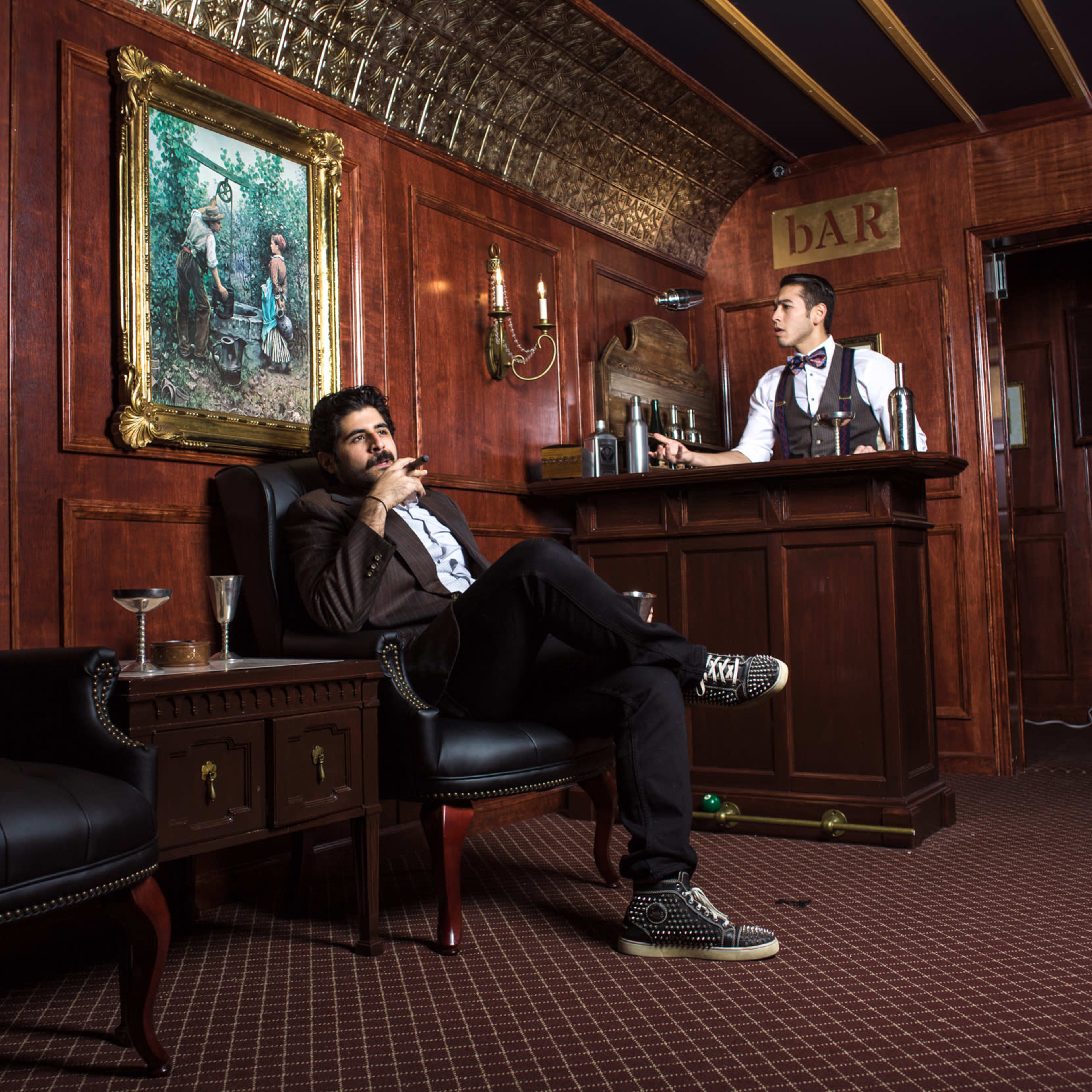 A man sits in an armchair smoking a cigar while another man stands behind a bar in a wooden-paneled room adorned with a large painting.
