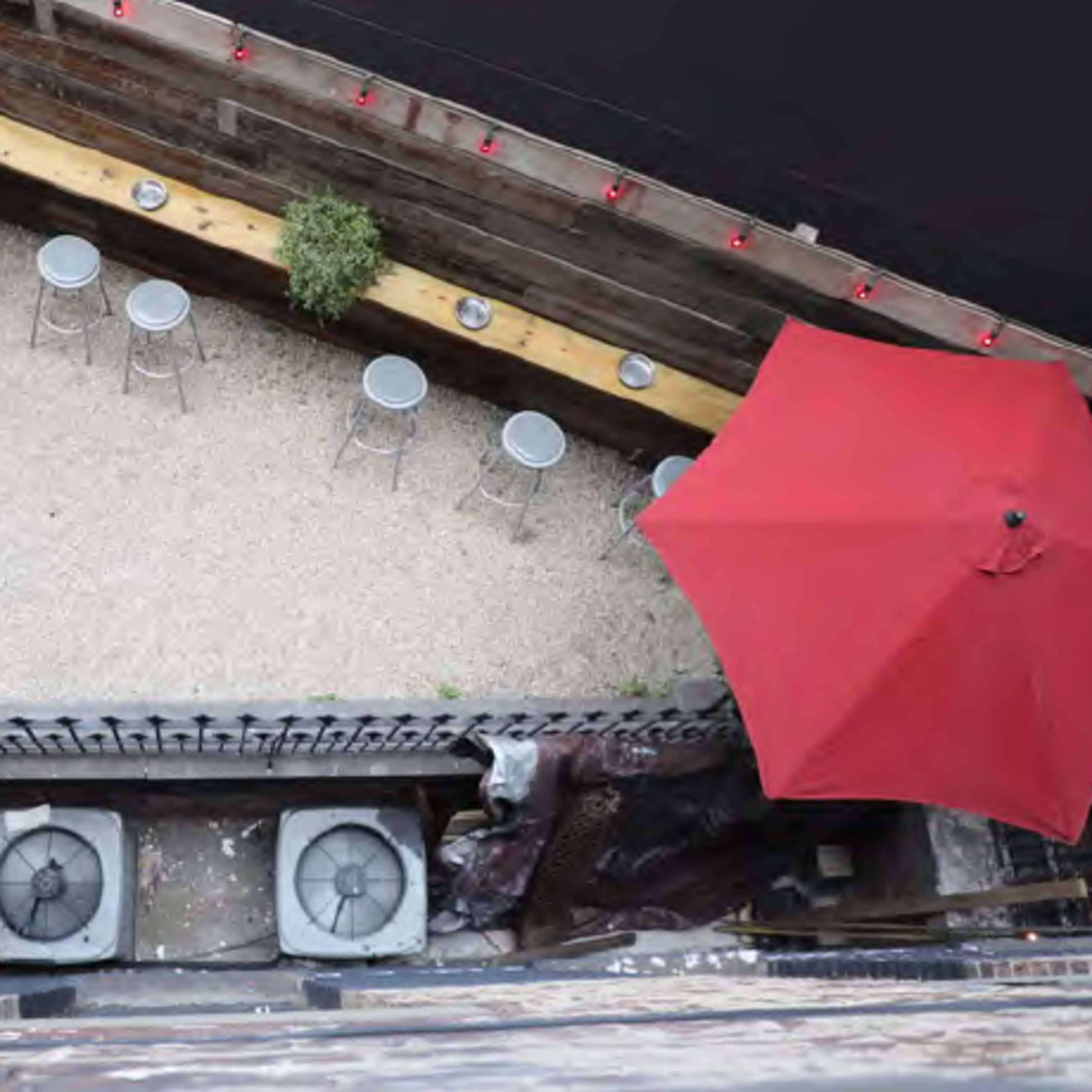 A narrow outdoor space with a gravel floor, featuring a long wooden table with stools and an umbrella.