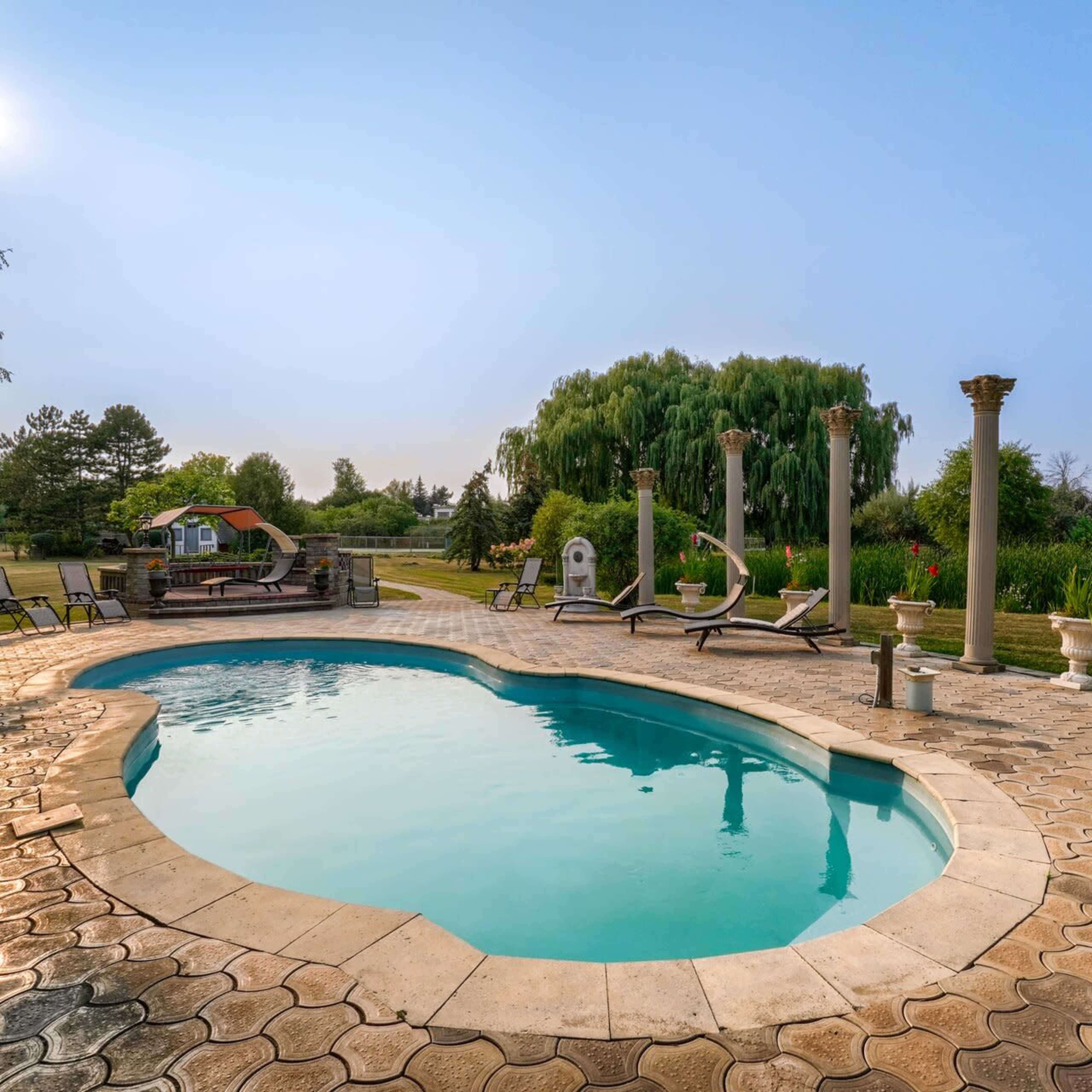 A swimming pool surrounded by stone decking and landscaped gardens, with several tall columns and patio furniture nearby.