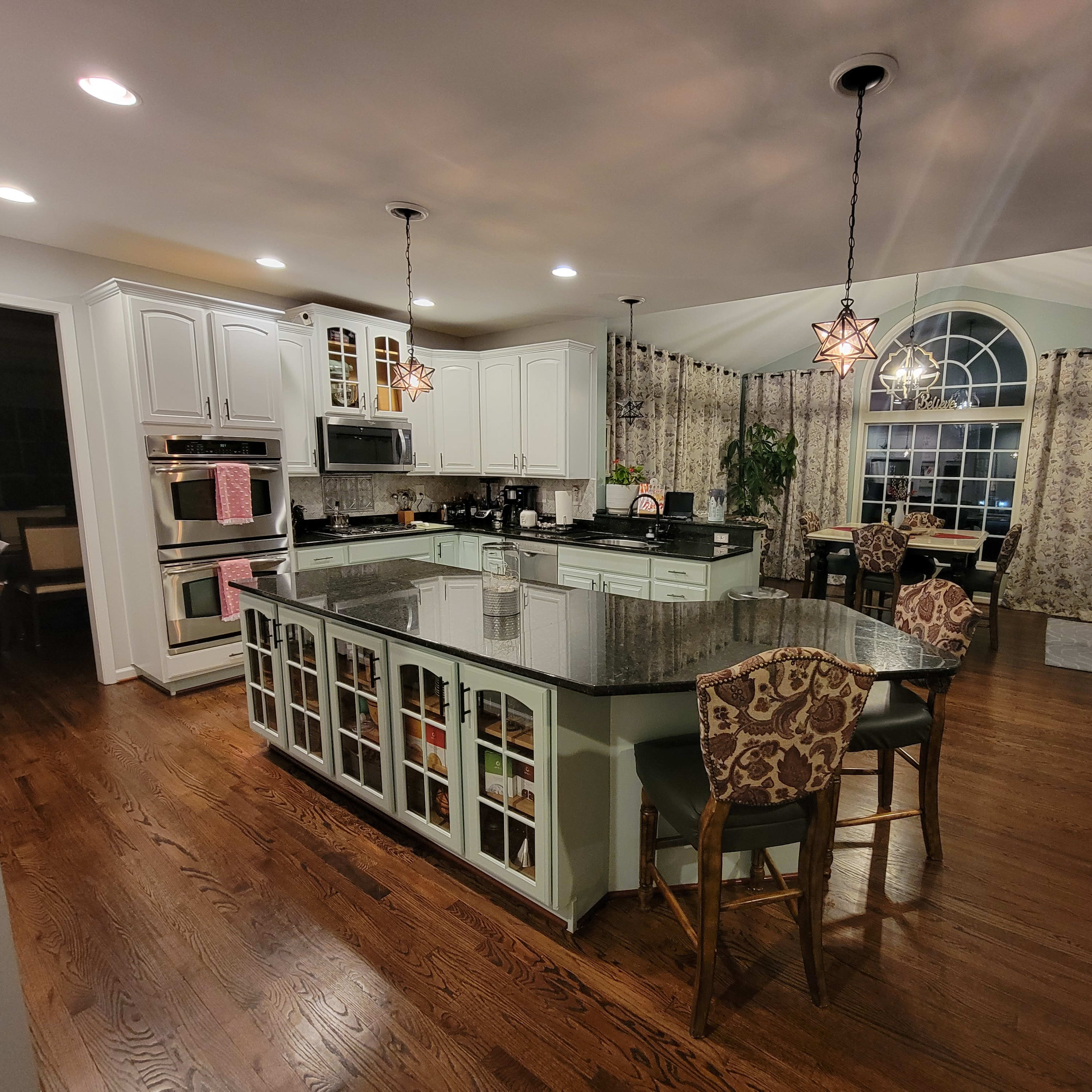 A spacious kitchen with a large island, dark wood flooring, white cabinetry, and a large window with ornate curtains.