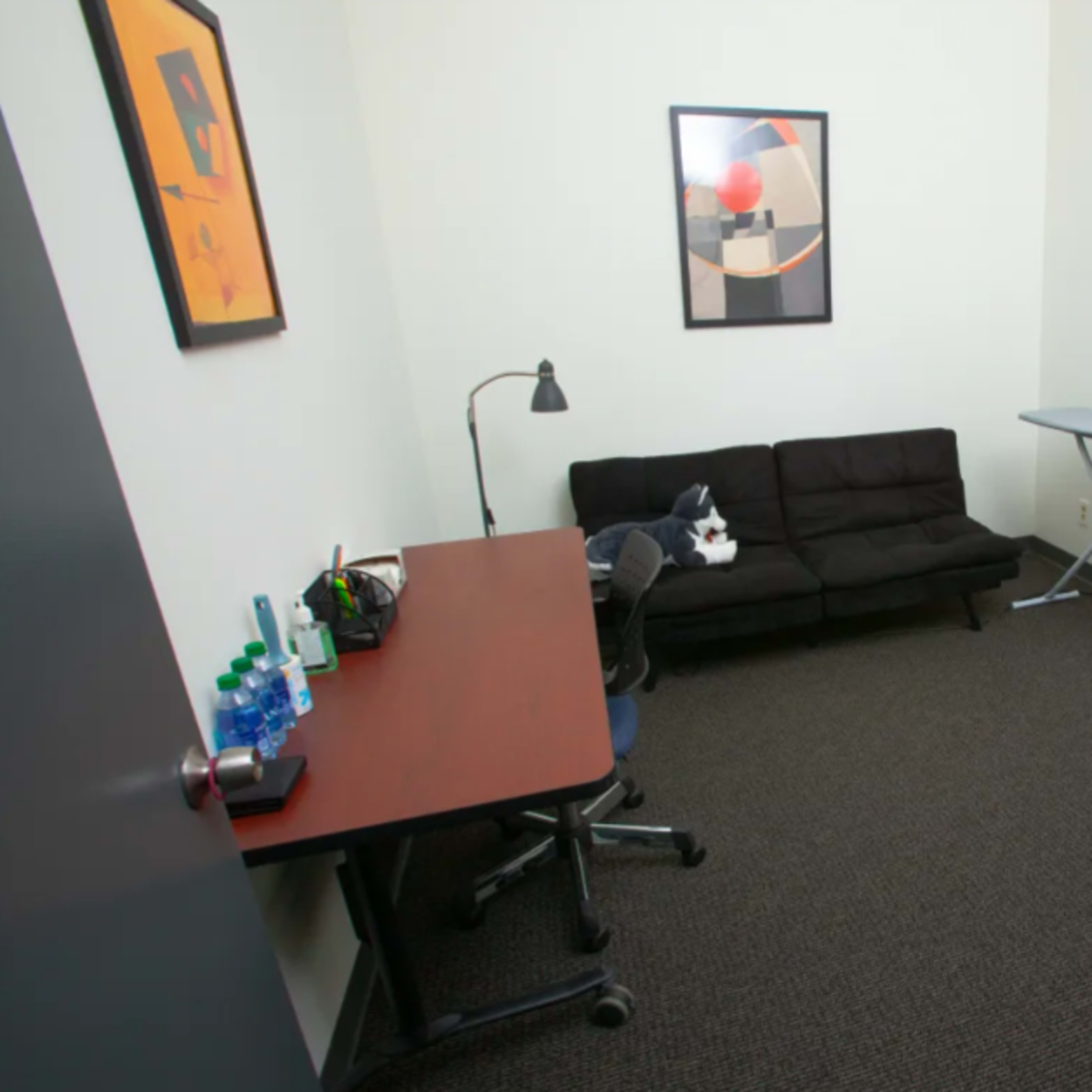 A small room with a black sofa, a desk with bottles, an ironing board, a mirror, and a blue armchair.