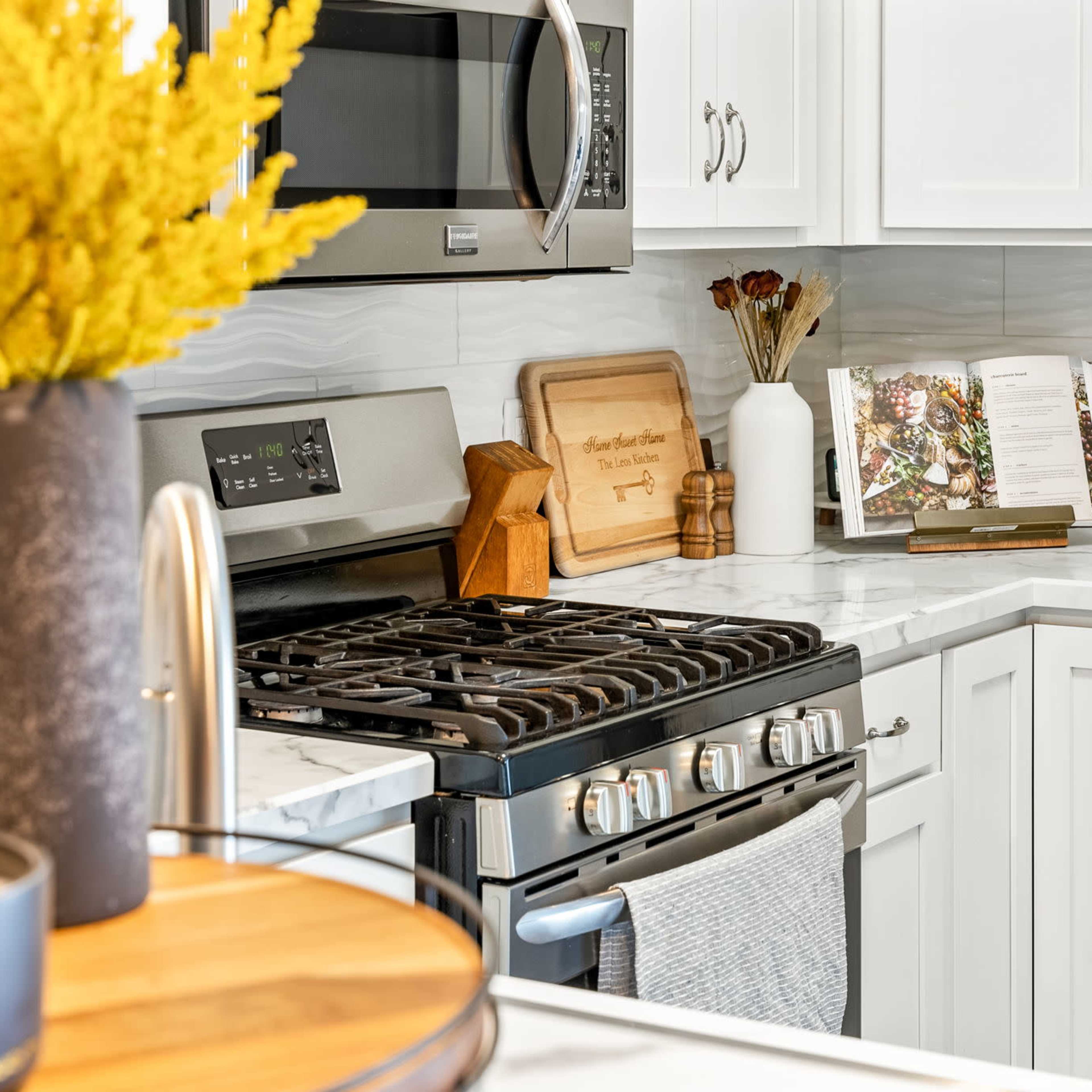 A modern kitchen features a stainless steel microwave, a gas stove with a light gray dish towel, and white cabinetry, with decorative items including a flower arrangement and a recipe book on the countertop.