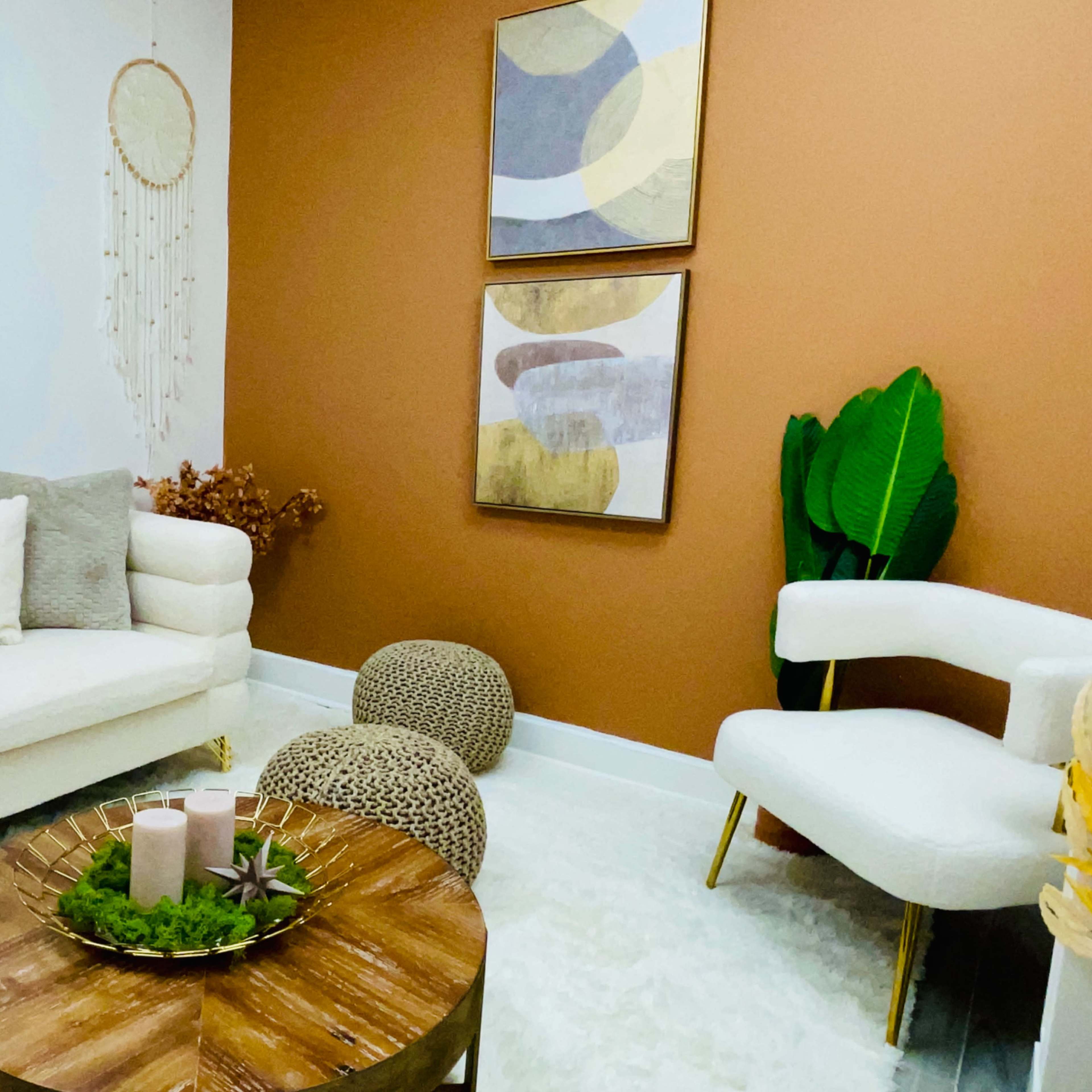 A cozy living room with a beige couch, two decorative paintings on a brown wall, and a wooden coffee table surrounded by plants and poufs.