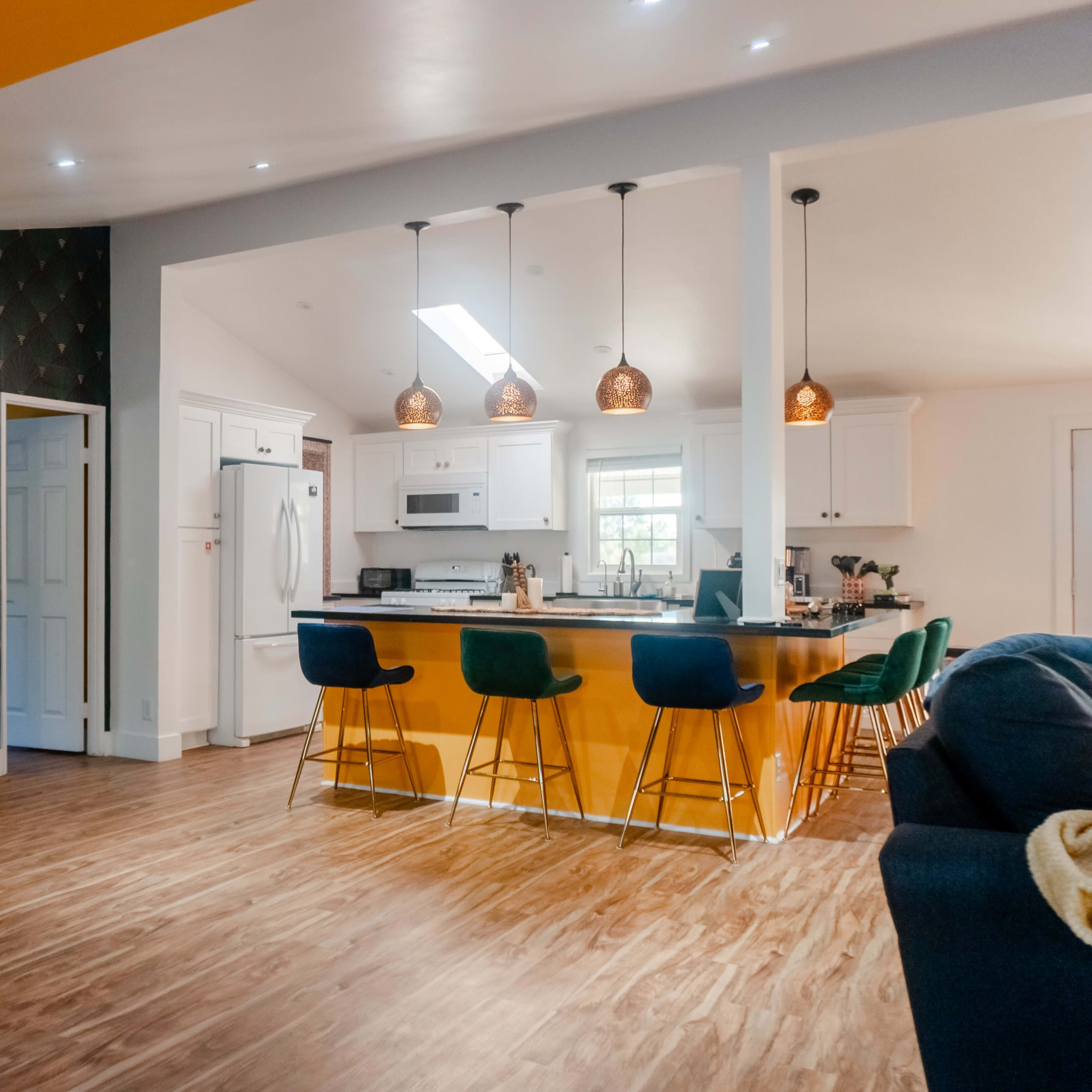 A modern kitchen area features a yellow accent wall, pendant lights, and a bar with colorful stools, alongside a cozy seating area with a wicker chair.