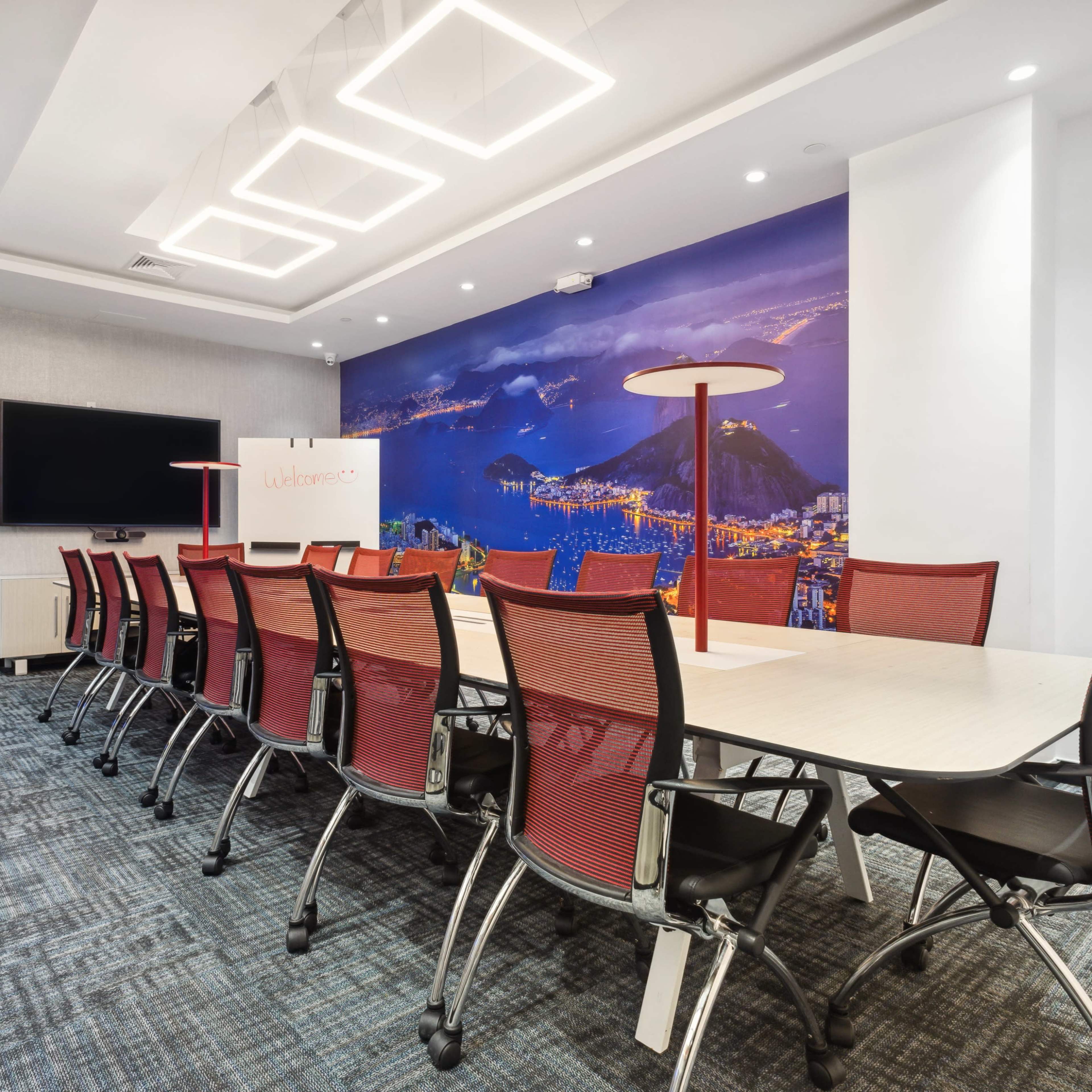 A modern conference room features a long table with red mesh chairs, a large screen on the wall, and a cityscape mural in the background.