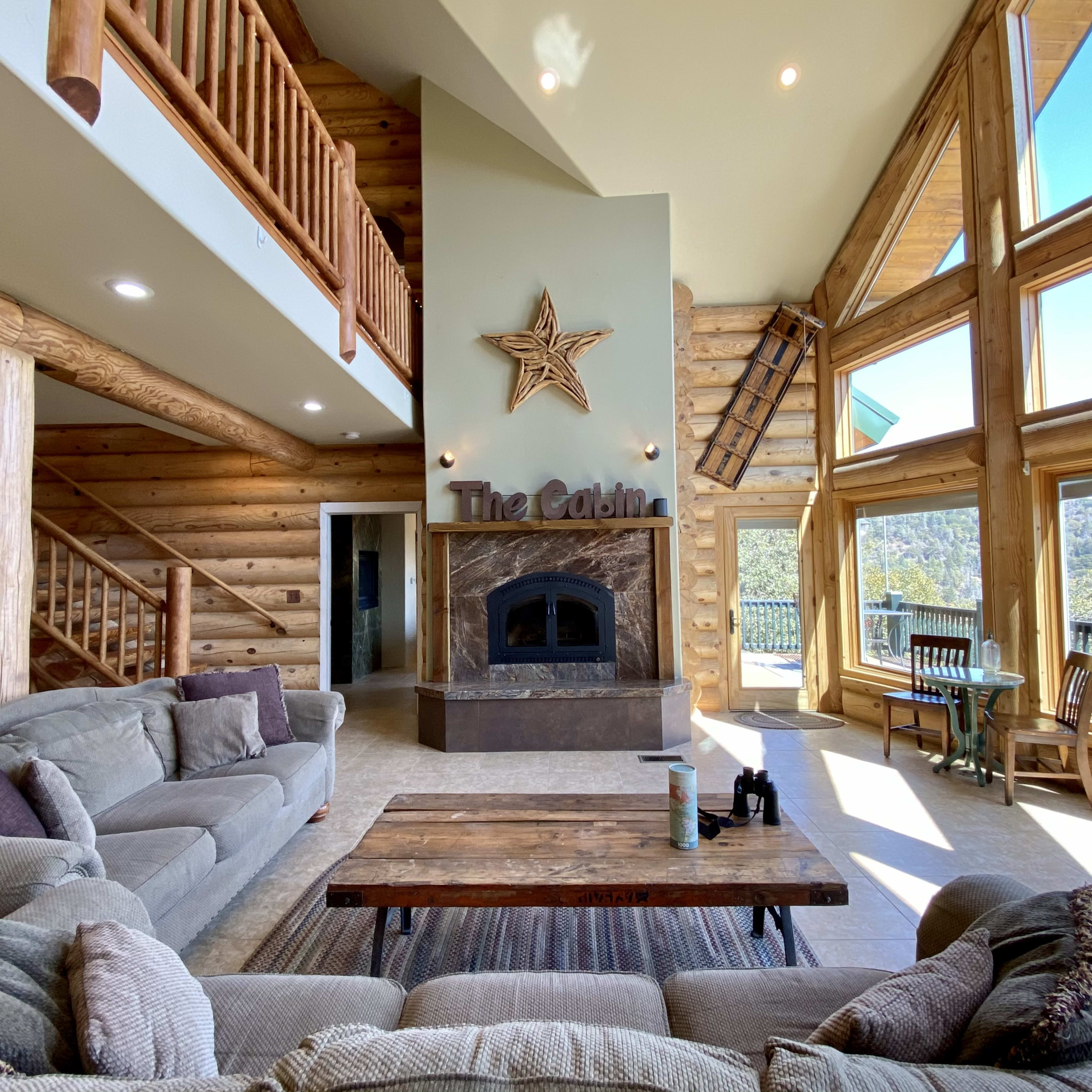 A spacious cabin interior with large windows, a stone fireplace, and wooden beams.