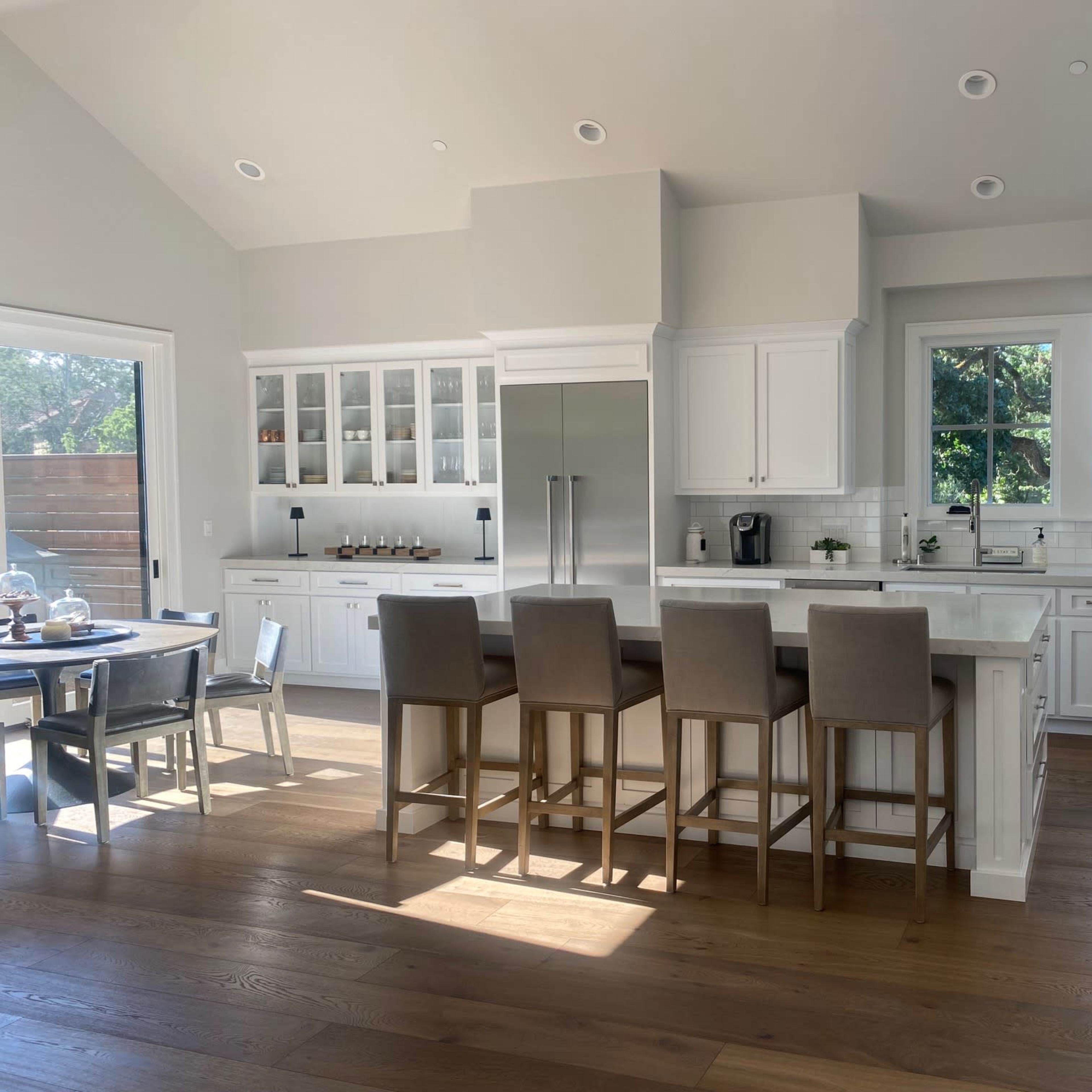 A modern kitchen with white cabinetry, a central island with bar stools, large windows, and a dining table set for meals.