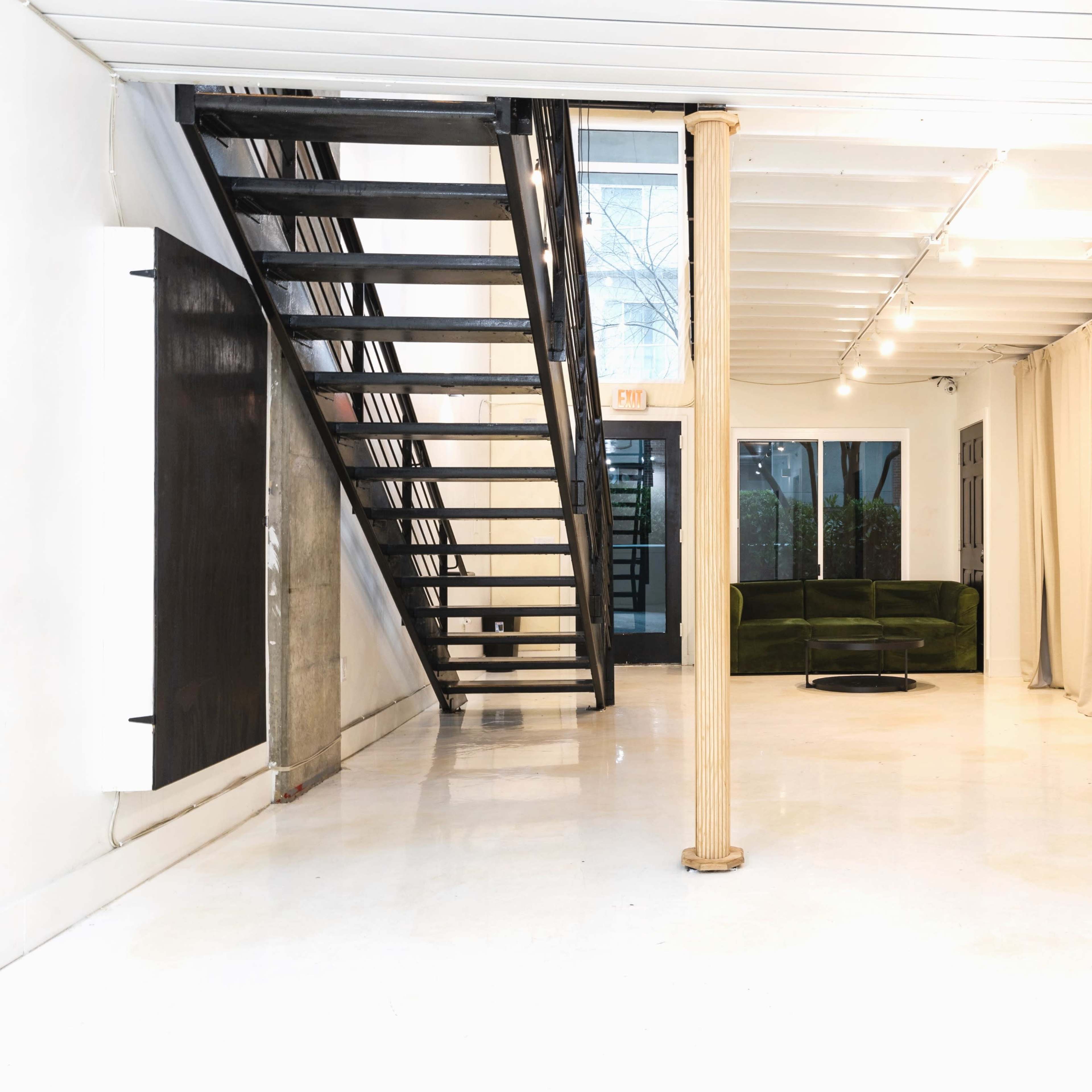 A modern interior space featuring a staircase on the left, a green sofa, and an open layout with polished floors and large windows.