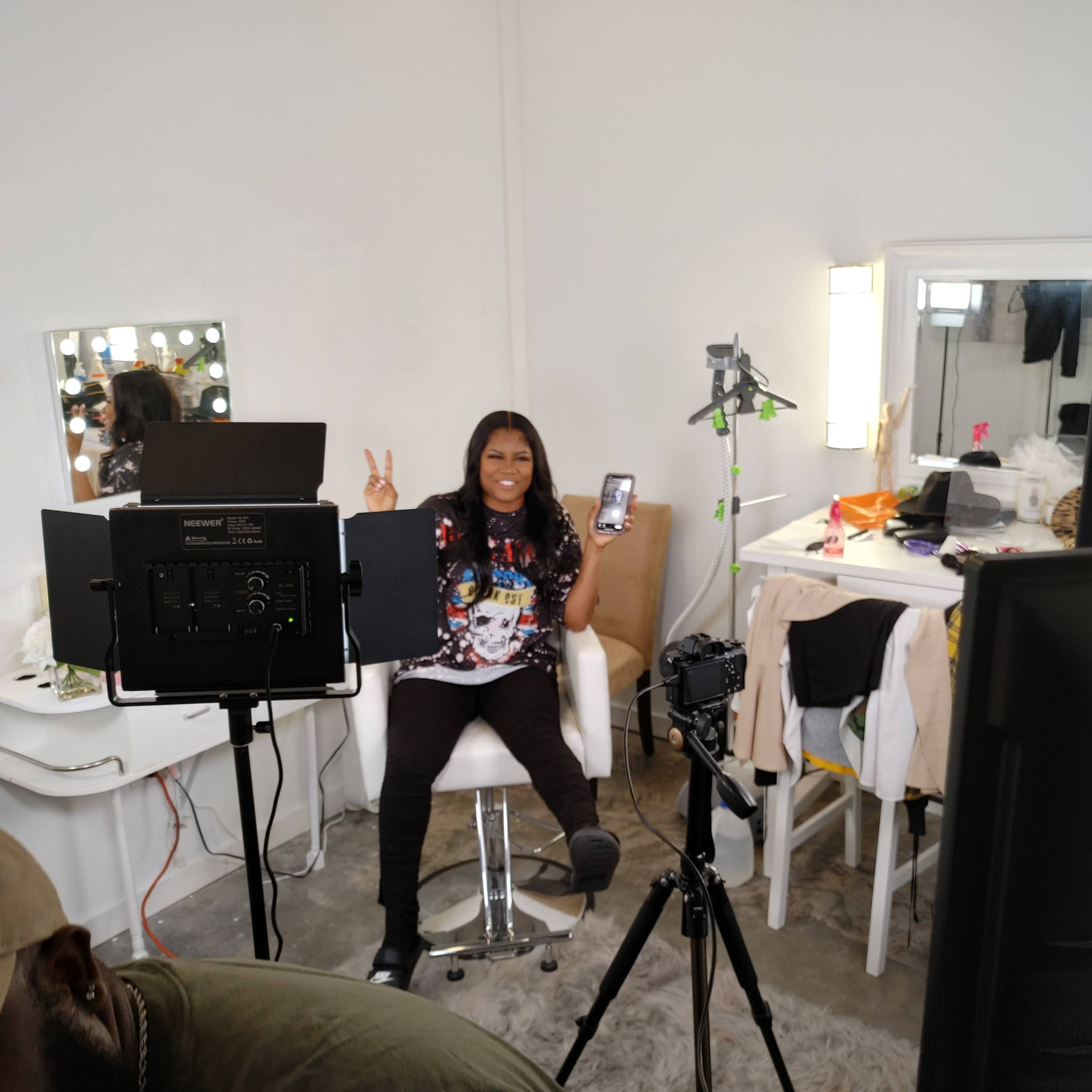 A person sits on a white chair in front of a makeup mirror, holding a phone and making a peace sign, while cameras and lighting equipment are set up around them.