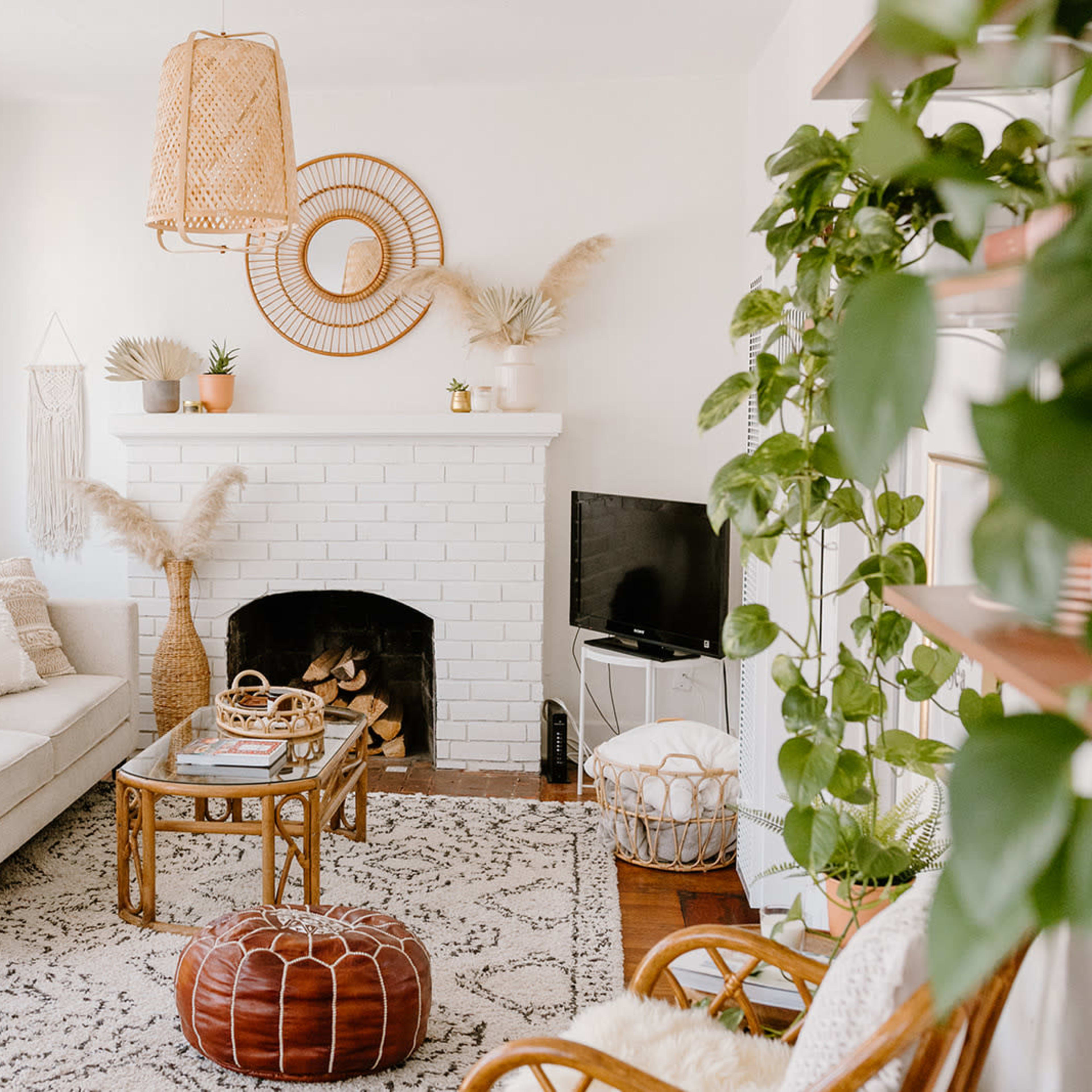 A cozy living room features a white brick fireplace, a light-colored sofa, a round mirror above the mantel, and a variety of plants throughout the space.