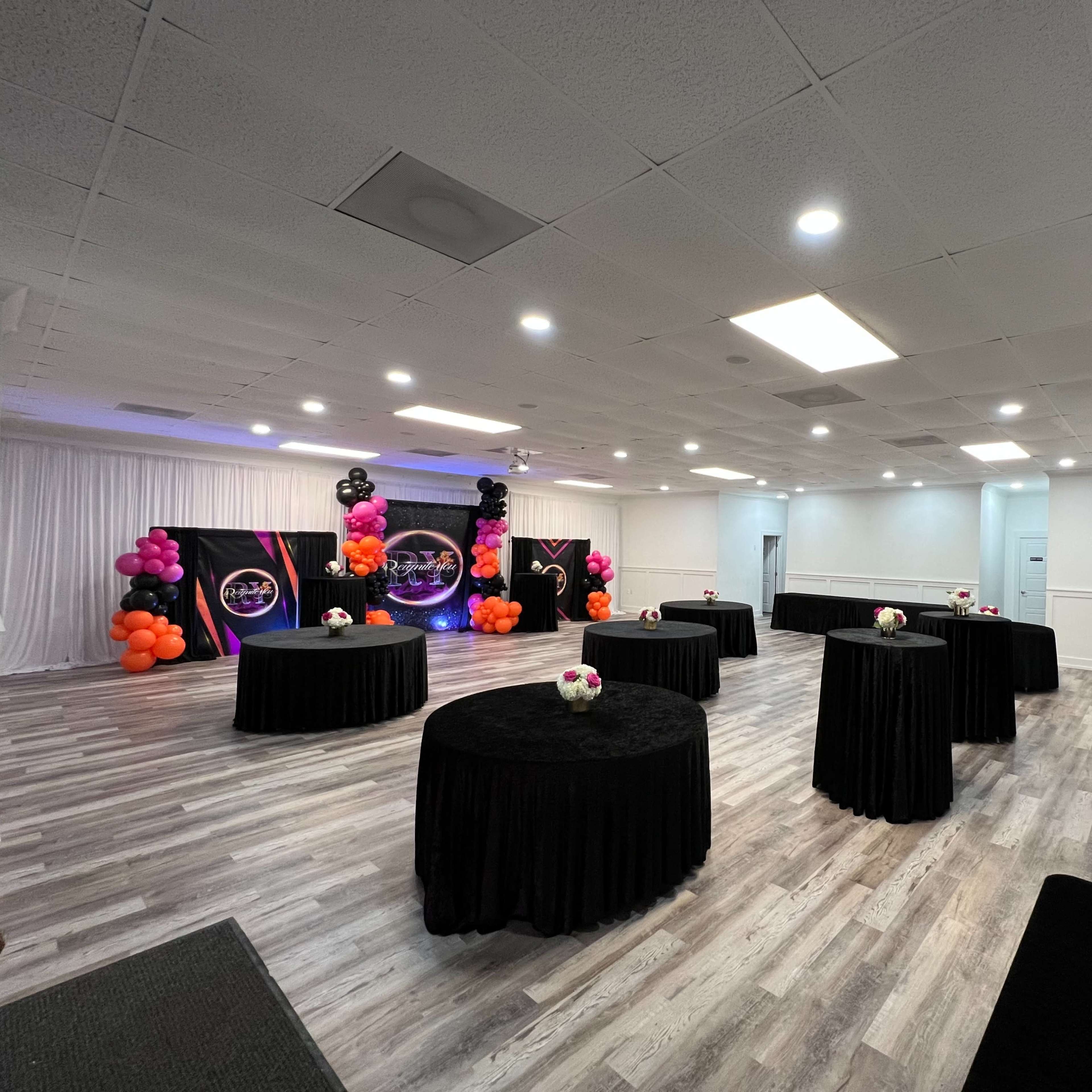 A large event space with several round tables covered in black cloth and decorated with floral arrangements, surrounded by colorful balloon displays.