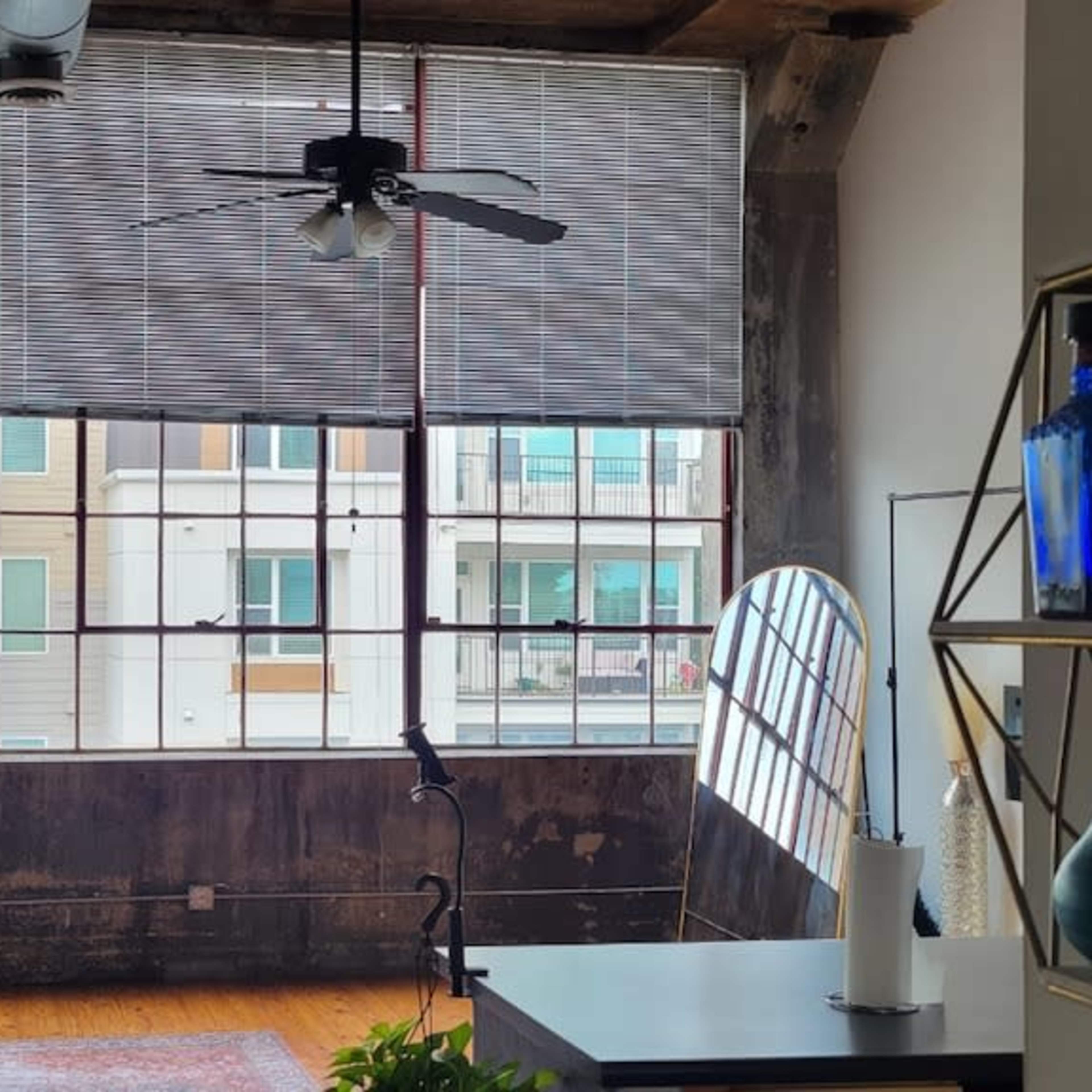 The scene depicts a modern living room with large windows, a ceiling fan, a light gray sofa, and a decorative shelving unit.