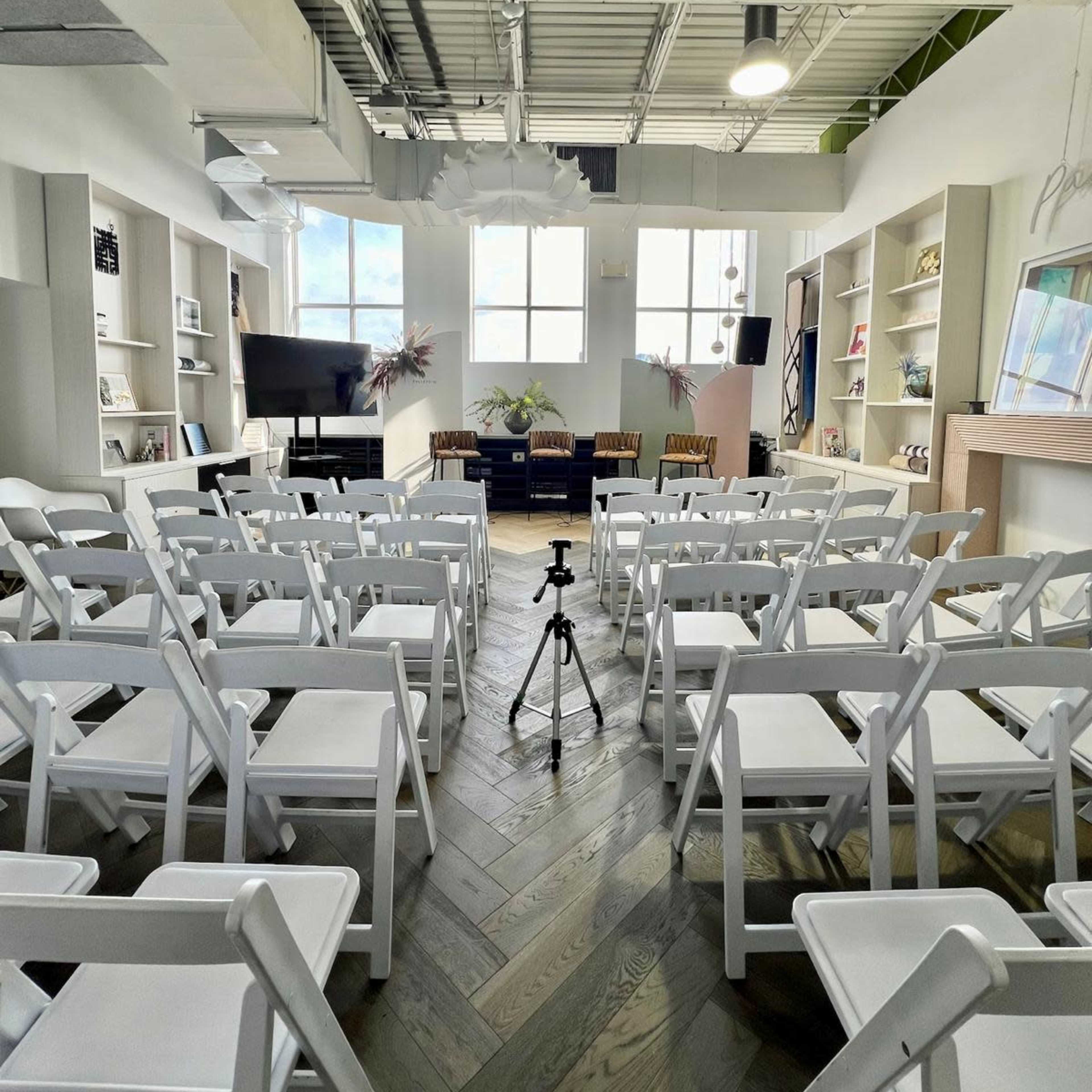 A spacious room is set up with white chairs arranged in rows facing a small stage, flanked by shelves displaying various items.