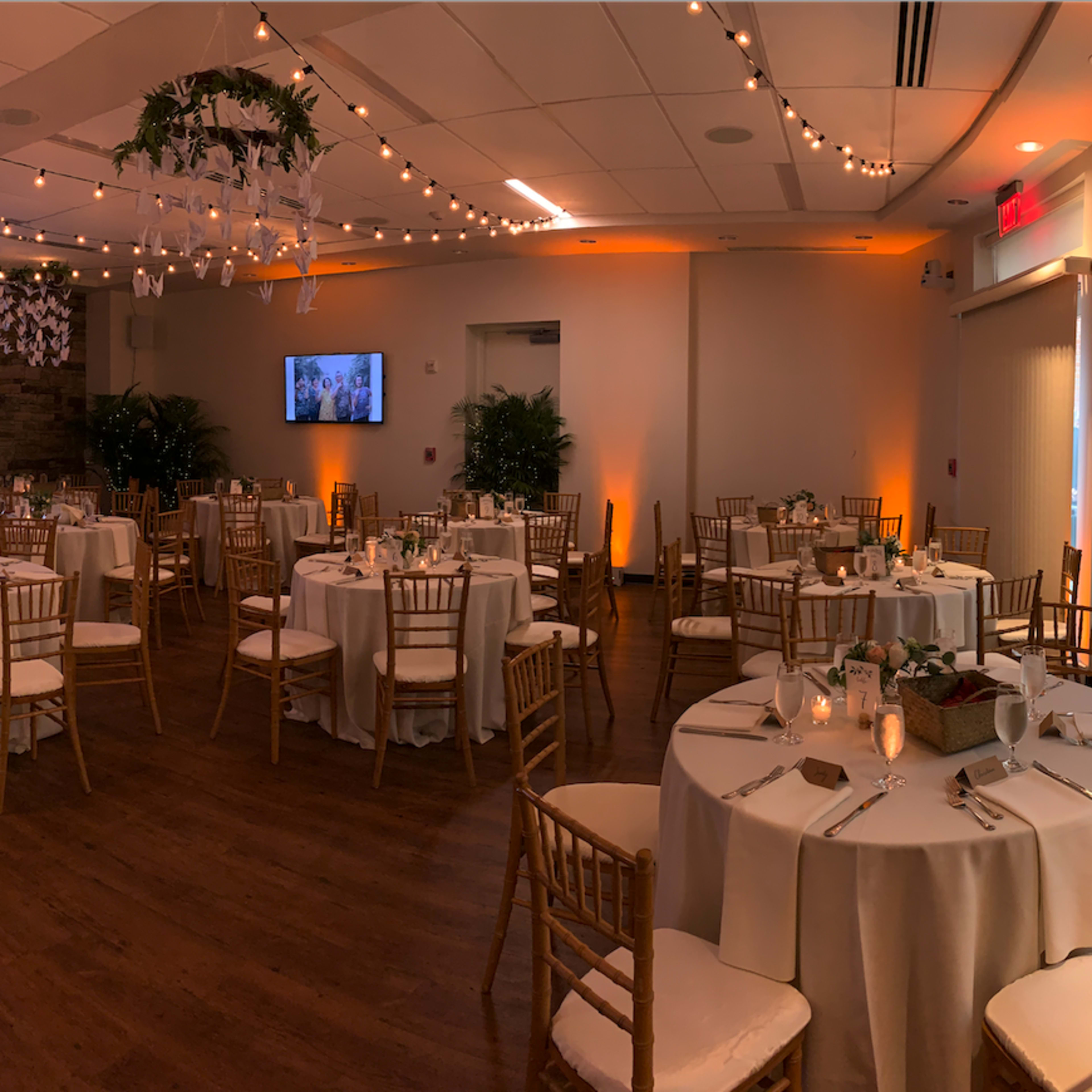 A well-decorated banquet hall with round tables set for an event, featuring a DJ booth and a large screen displaying photos.