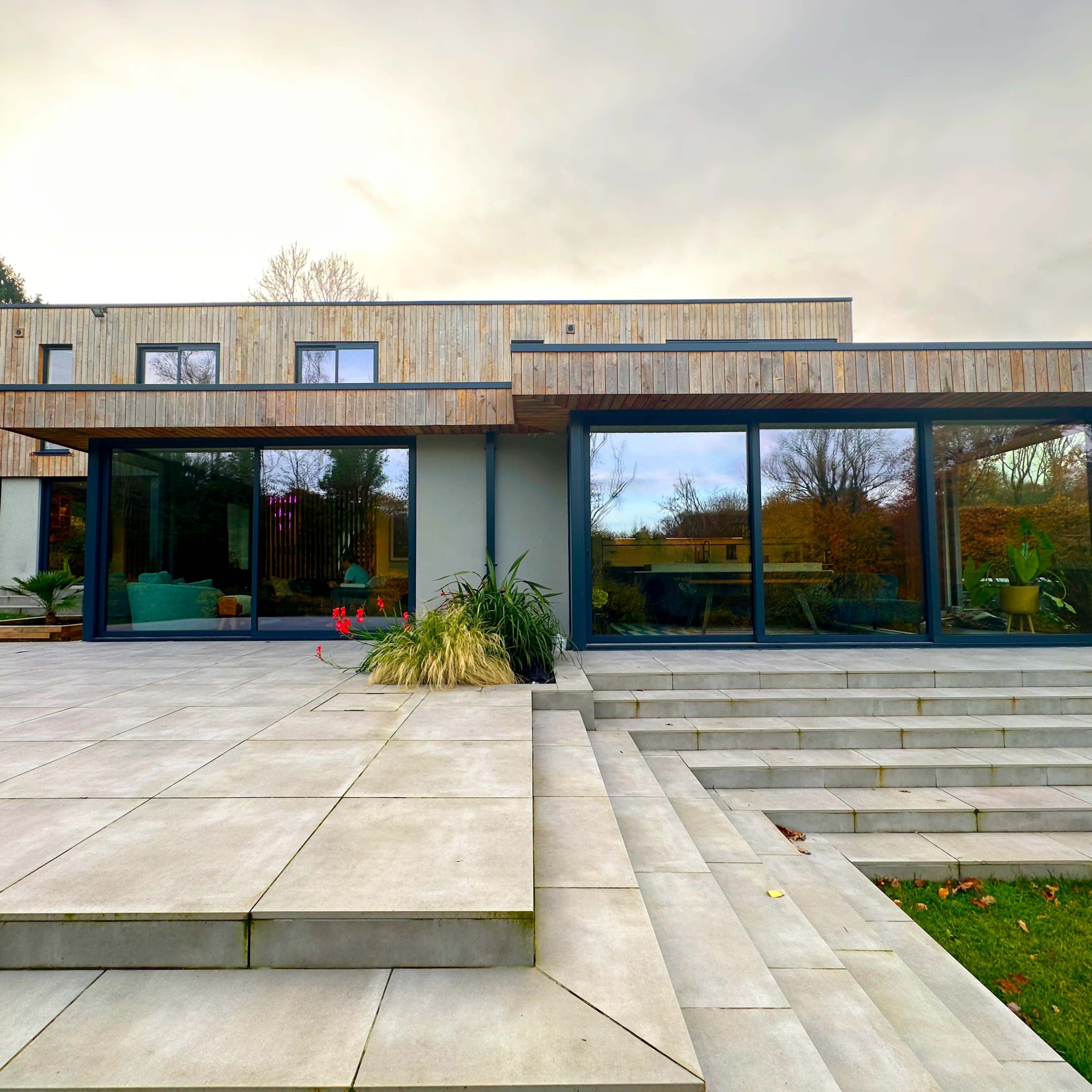 A modern two-story house with a wooden facade, large windows, and a paved patio leading to a well-manicured lawn.