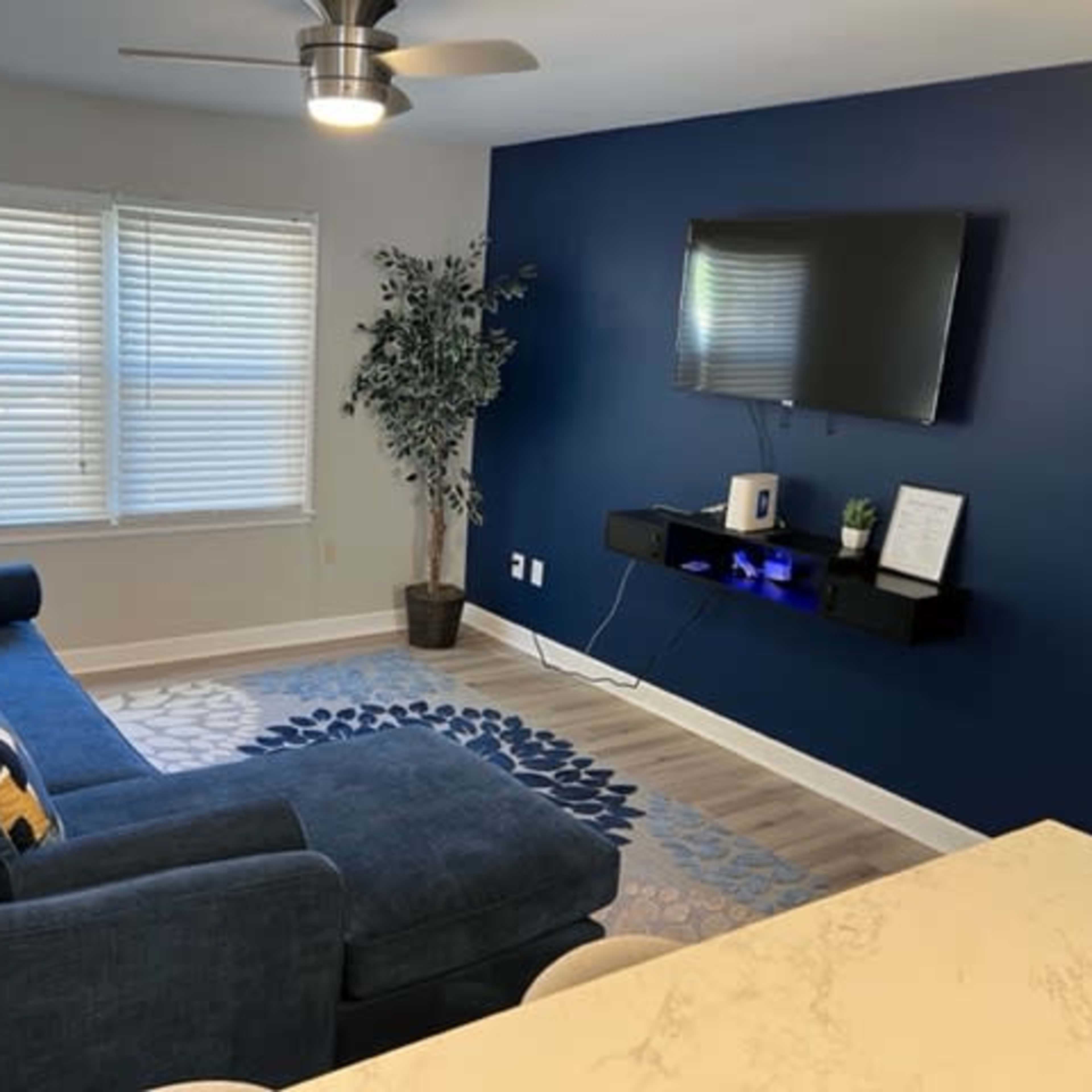 A living room featuring a blue sectional sofa, a wall-mounted TV, and a small shelf with decorative items against a dark blue wall.