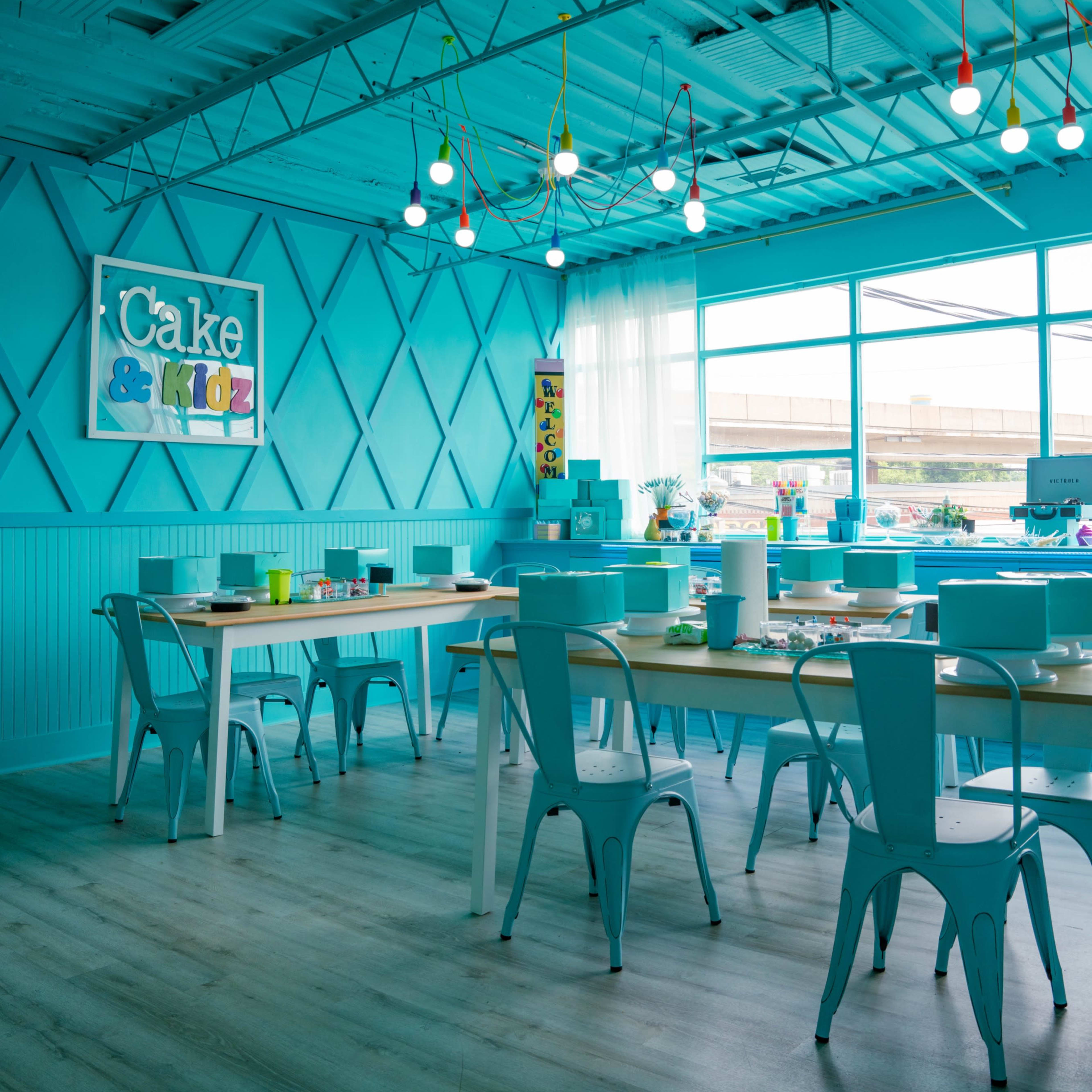 A brightly colored turquoise room with tables and chairs arranged for a baking or craft activity, featuring a sign that reads "Cake & Kids" on the wall.
