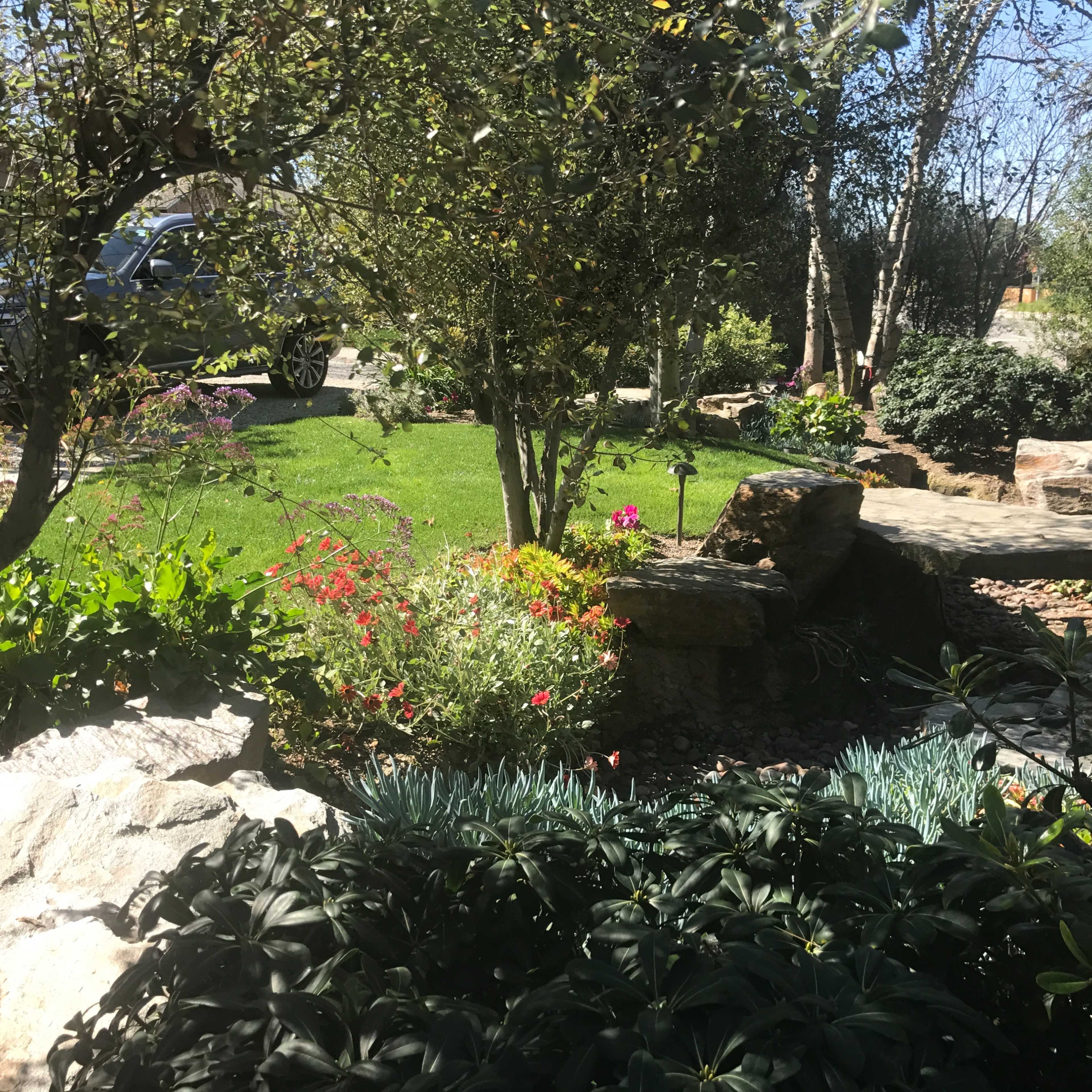 A well-kept garden features a variety of plants, flowers, and stone pathways alongside a neatly trimmed lawn and a parked car in the background.