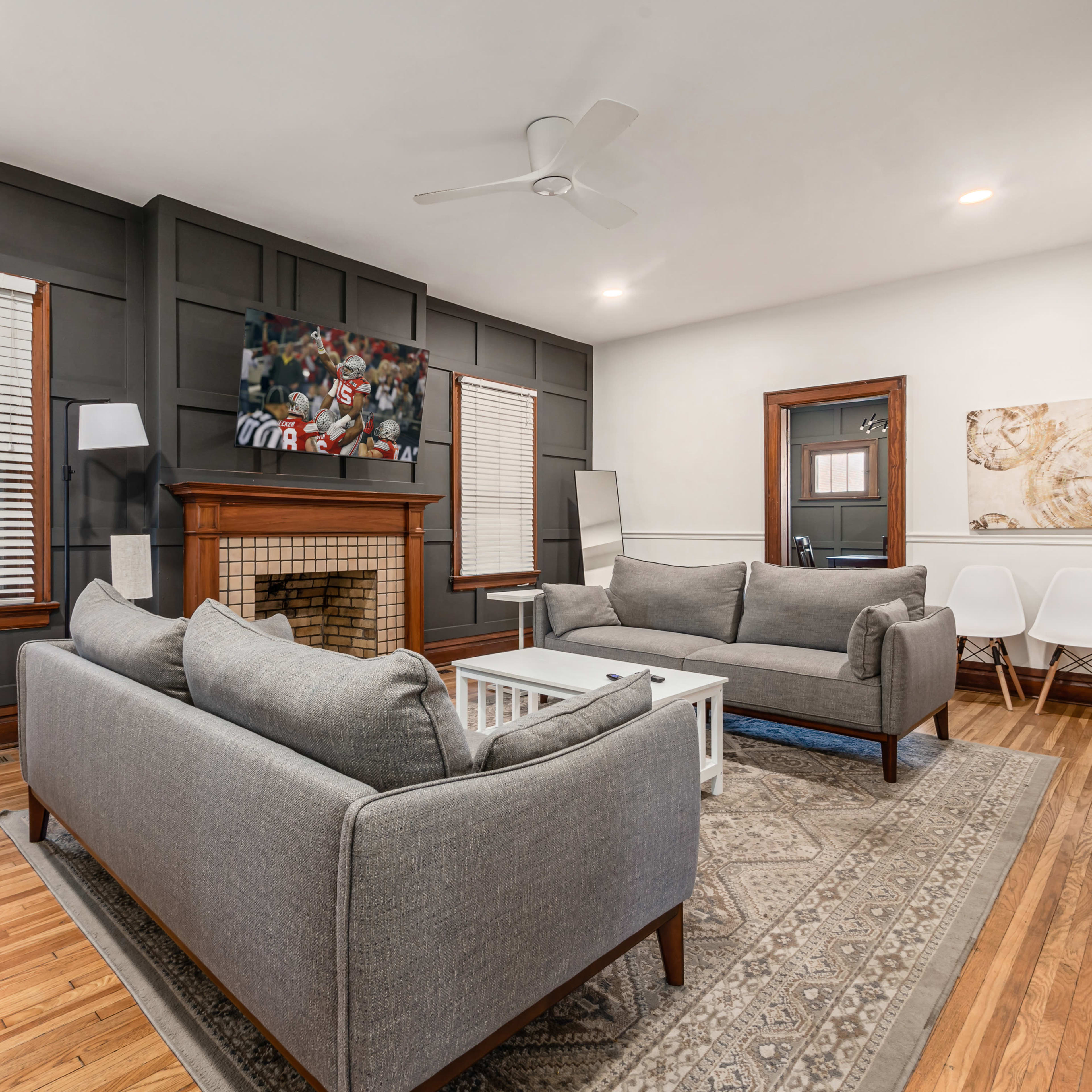 The living room features two gray sofas facing a wooden fireplace, a television mounted on the wall, and a coffee table on a patterned rug.