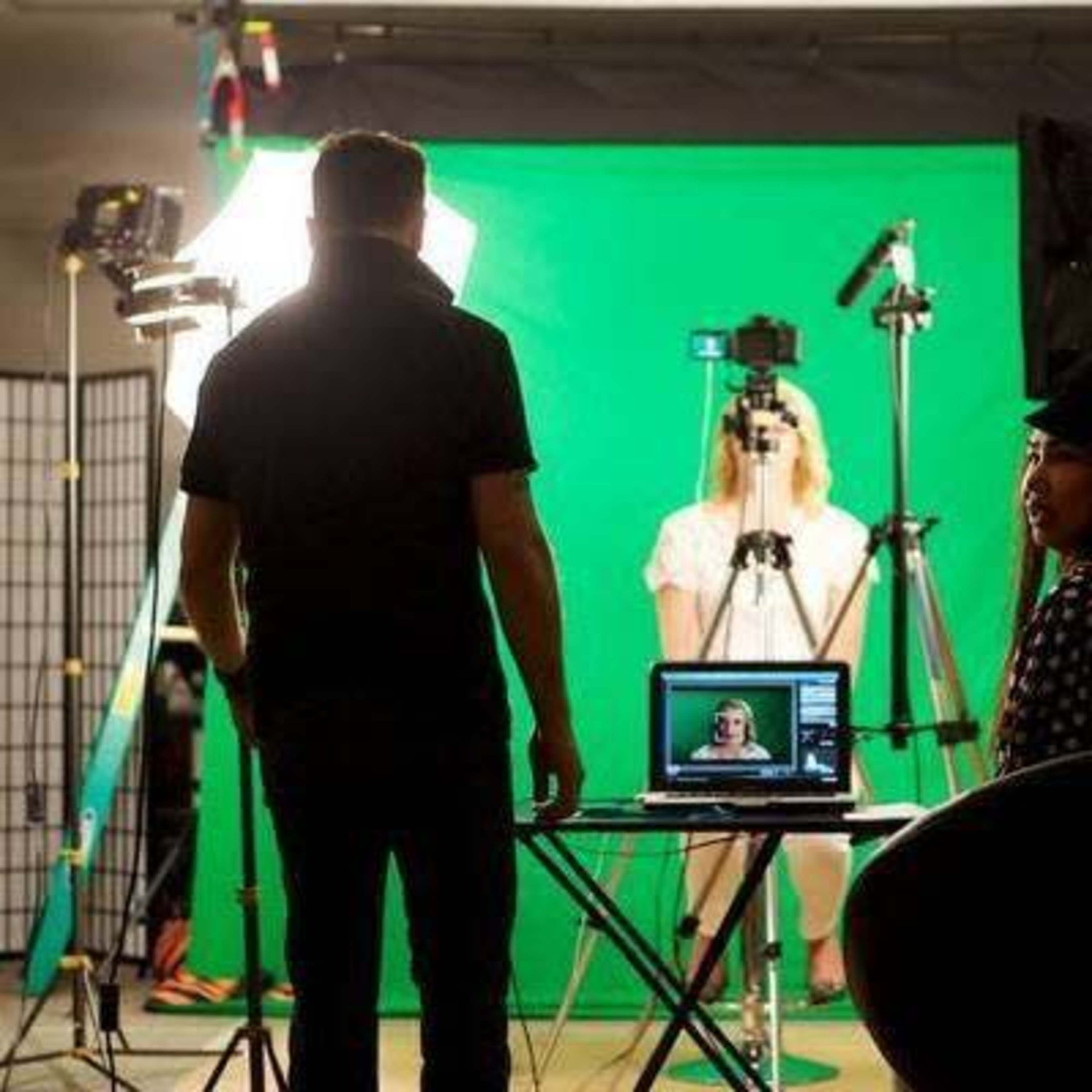 A person stands in front of a computer and camera setup, facing a subject positioned in front of a green screen.