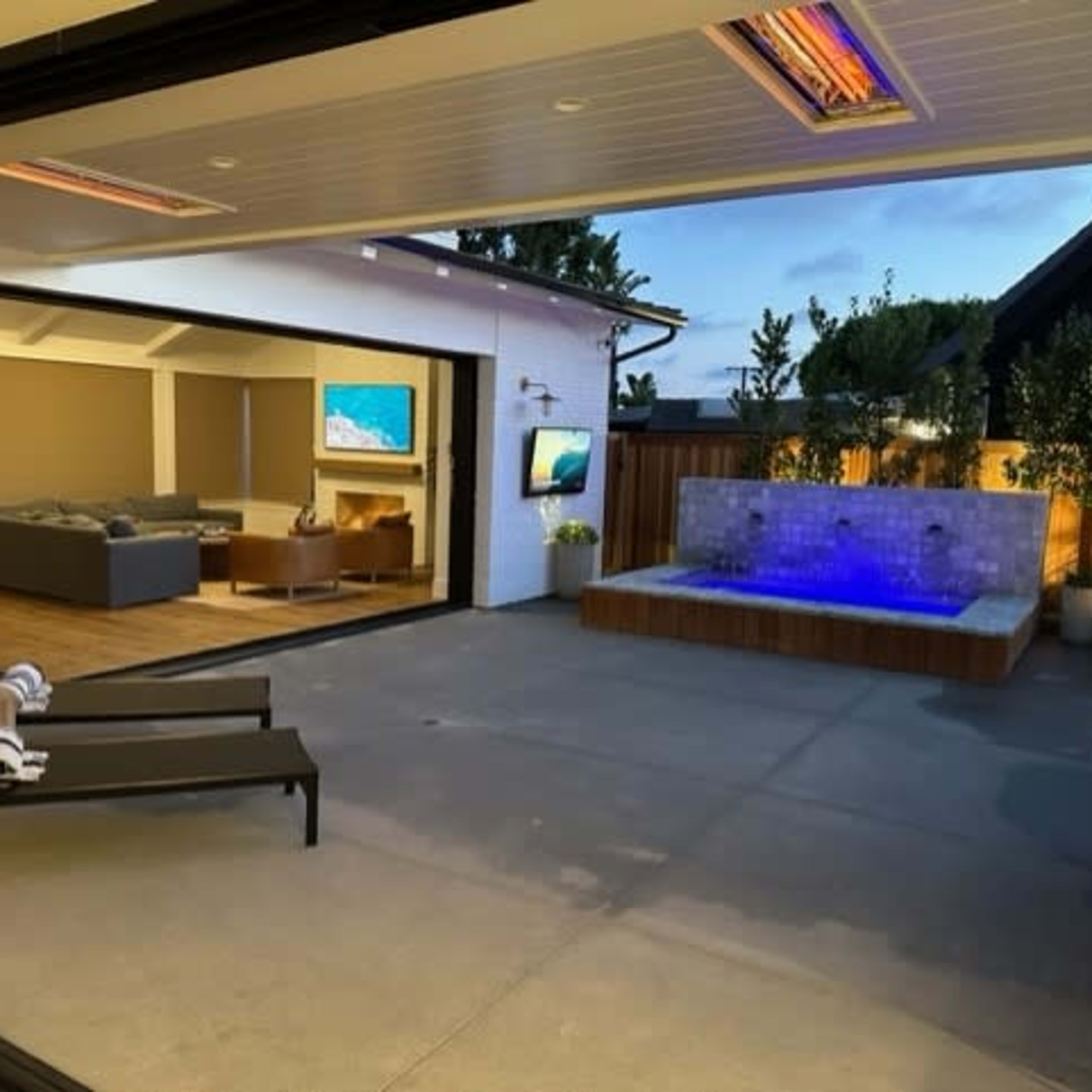 A modern outdoor space features a hot tub with blue illumination, a seating area with a gray couch, and large sliding glass doors that open to the interior.