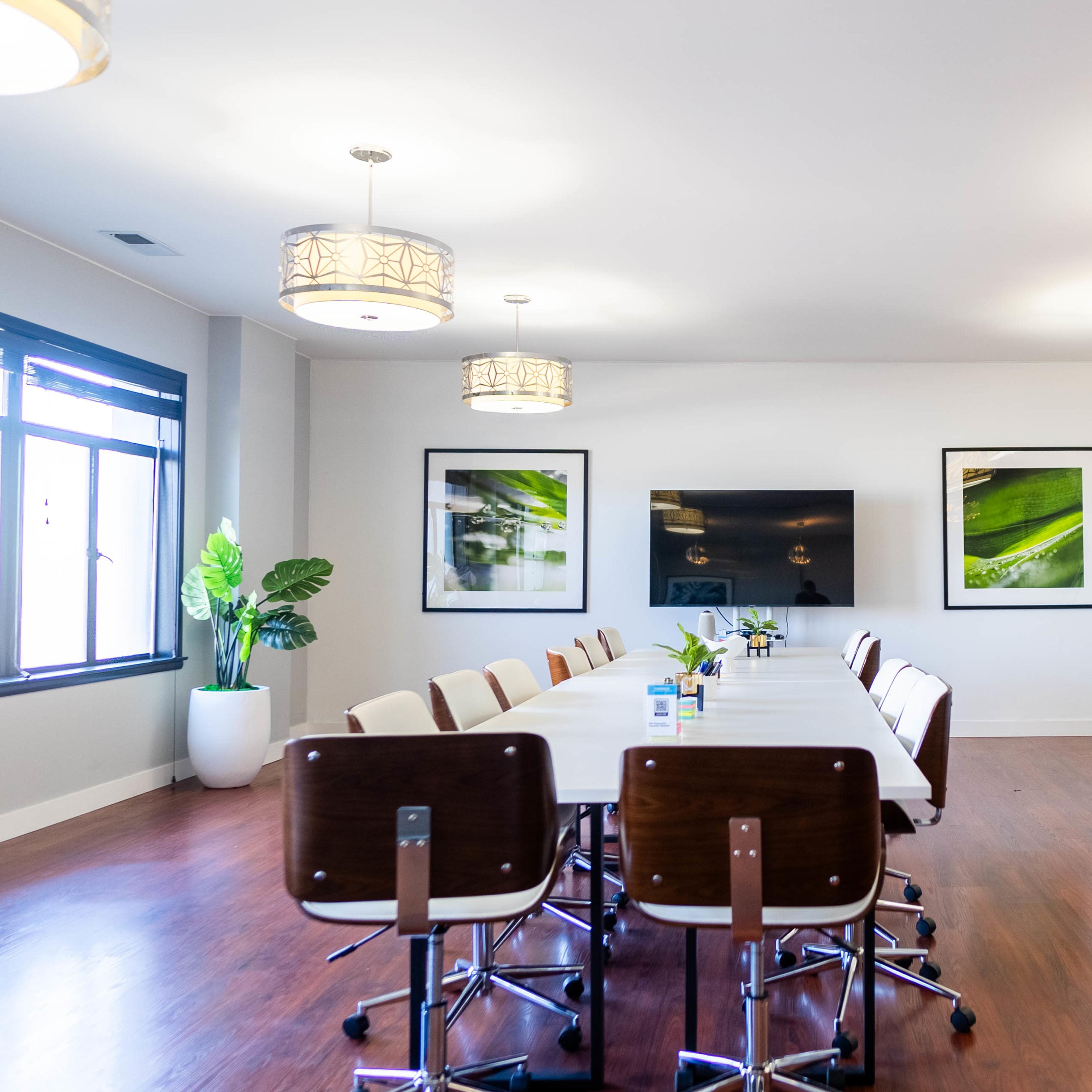 A modern conference room features a long table surrounded by chairs, with plants, artwork, and a large screen on the wall.