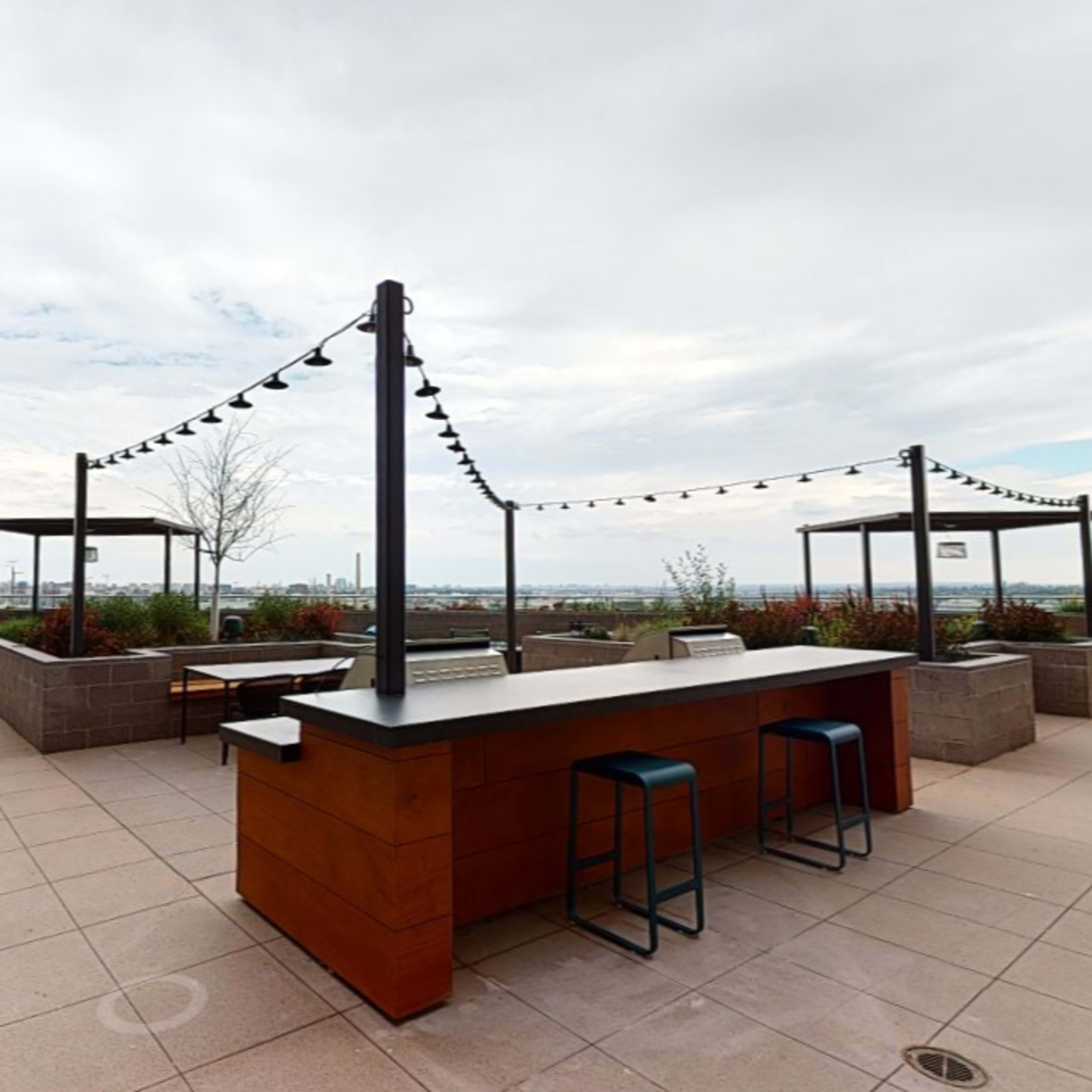 A rooftop patio area featuring a long bar with stools, surrounded by overhead lights and greenery, with a view of a city skyline in the distance.