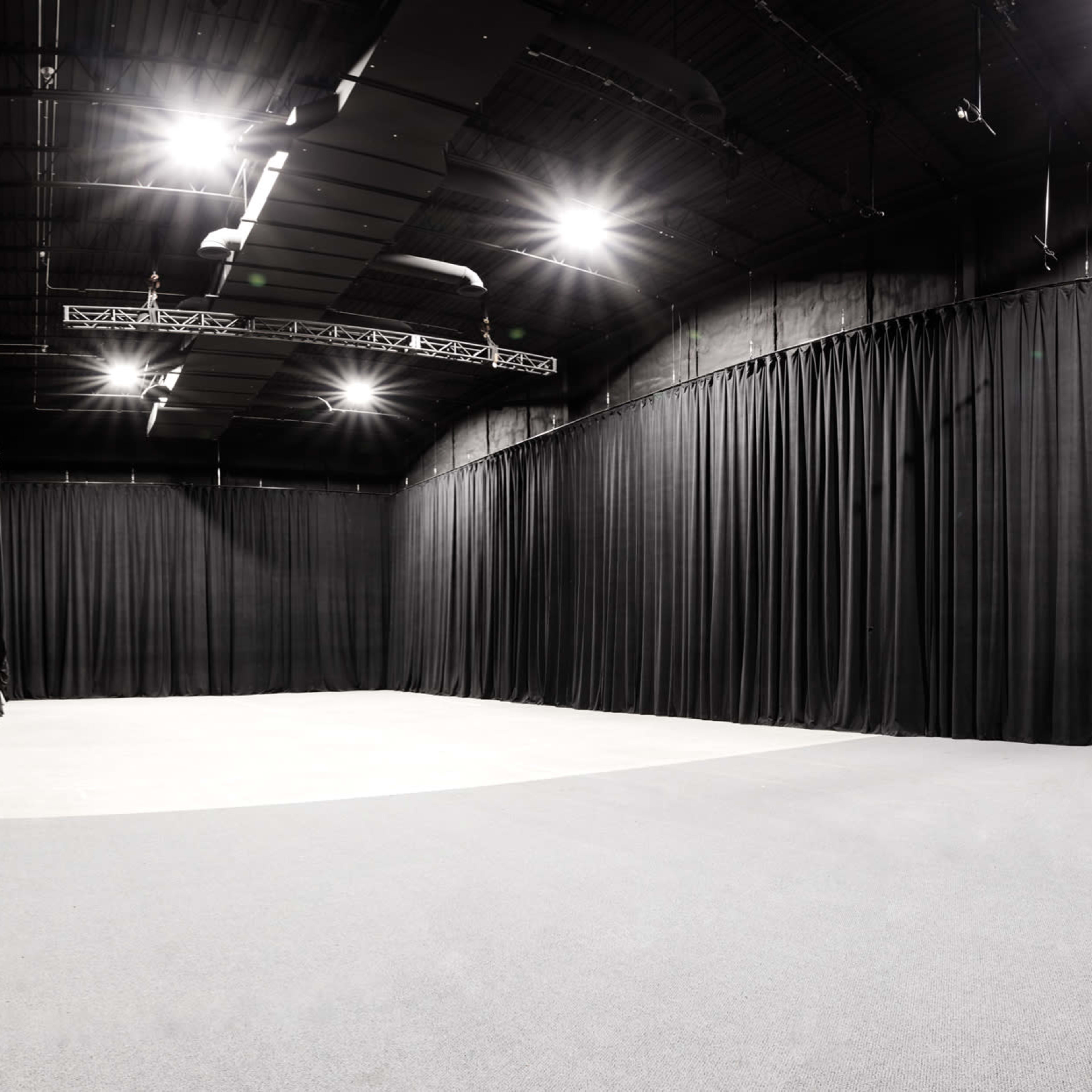 A spacious studio with black curtains lining the walls and bright overhead lights illuminating the gray floor.