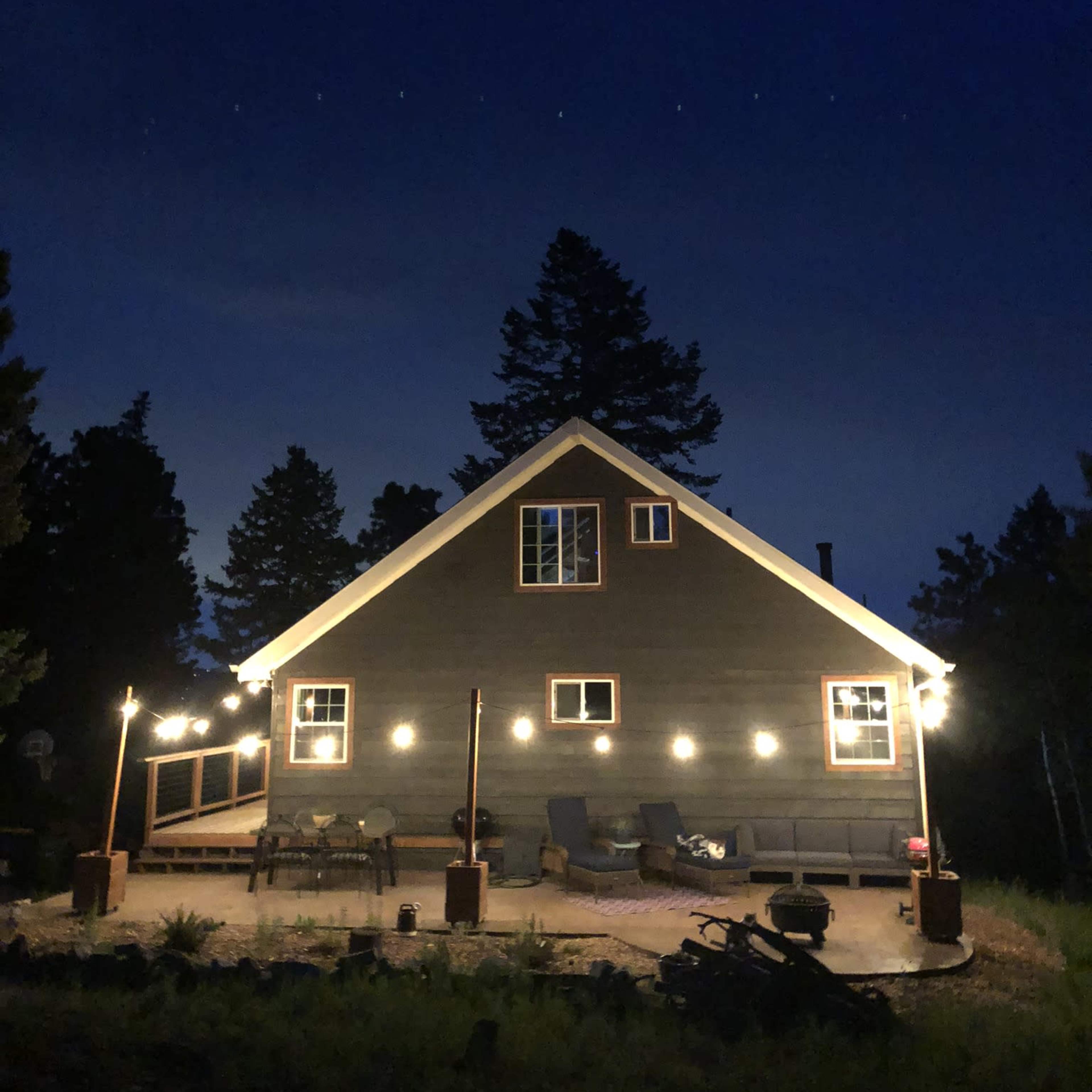 A dimly lit house with string lights illuminates its exterior against a dark night sky, surrounded by trees.
