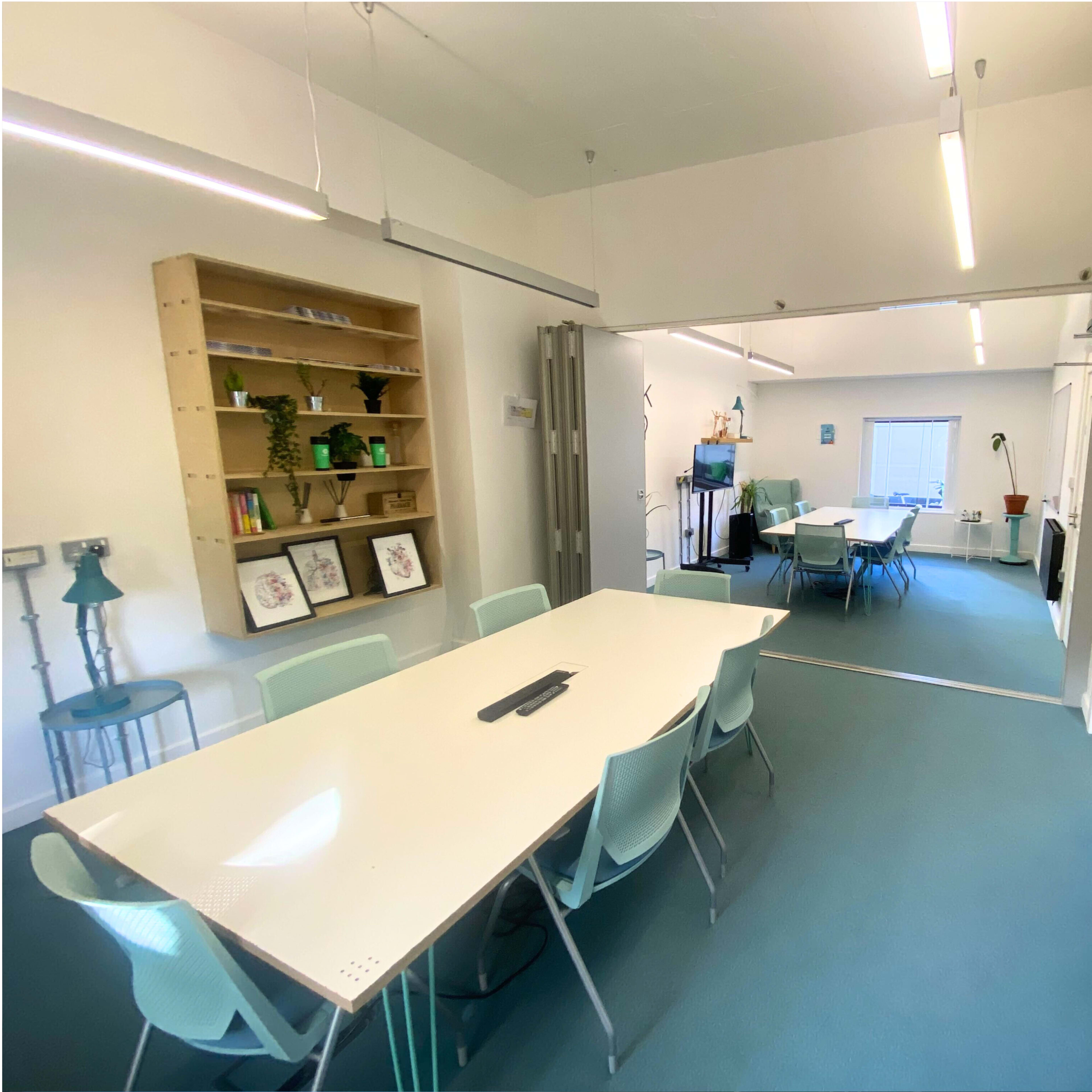 A modern meeting room with a large white table and light blue chairs, along with bookshelves and a whiteboard.