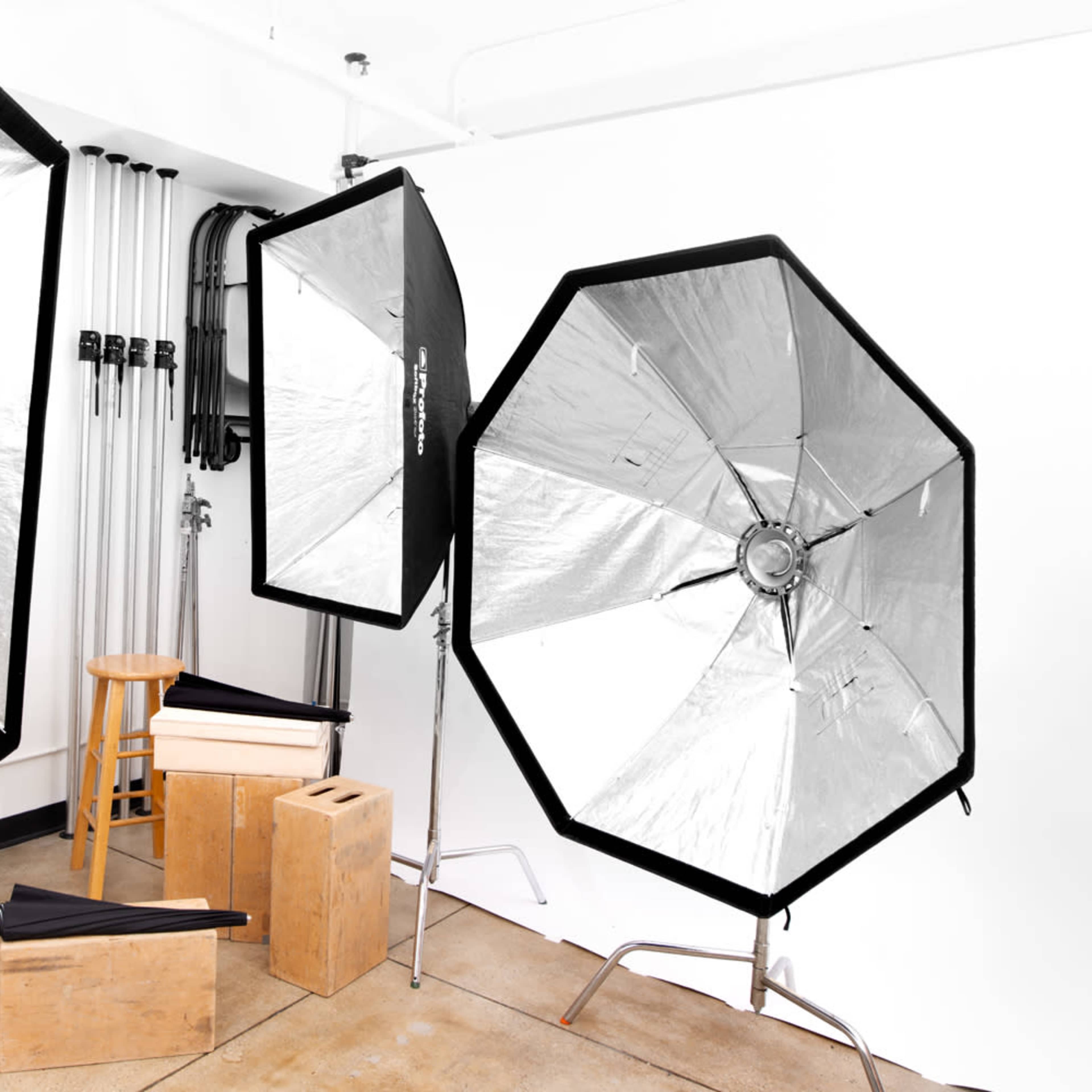 A photography setup with three light modifiers, a stool, and various wooden boxes against a white wall.