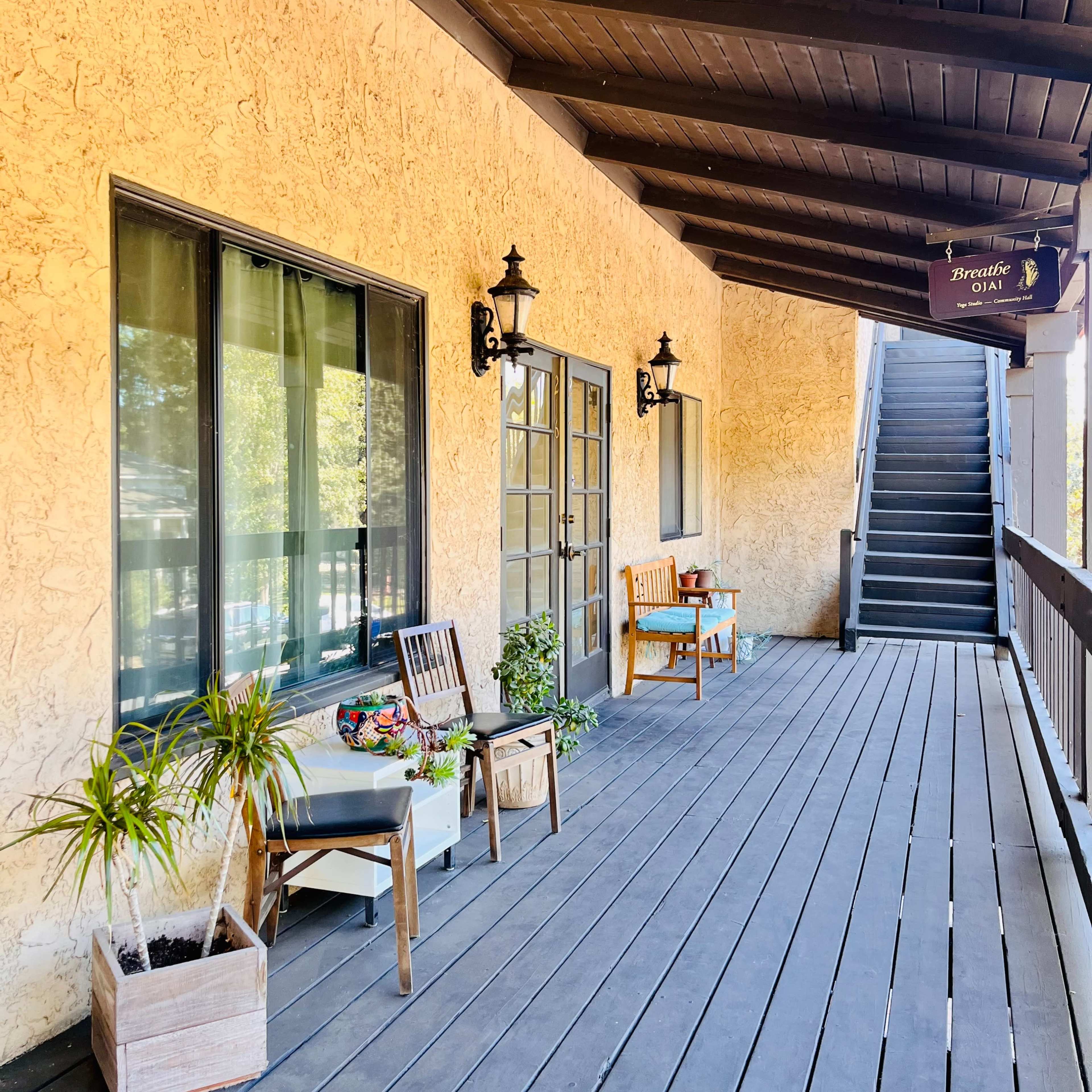 A covered wooden walkway with two chairs and potted plants beside a pair of glass doors, leading to a staircase.