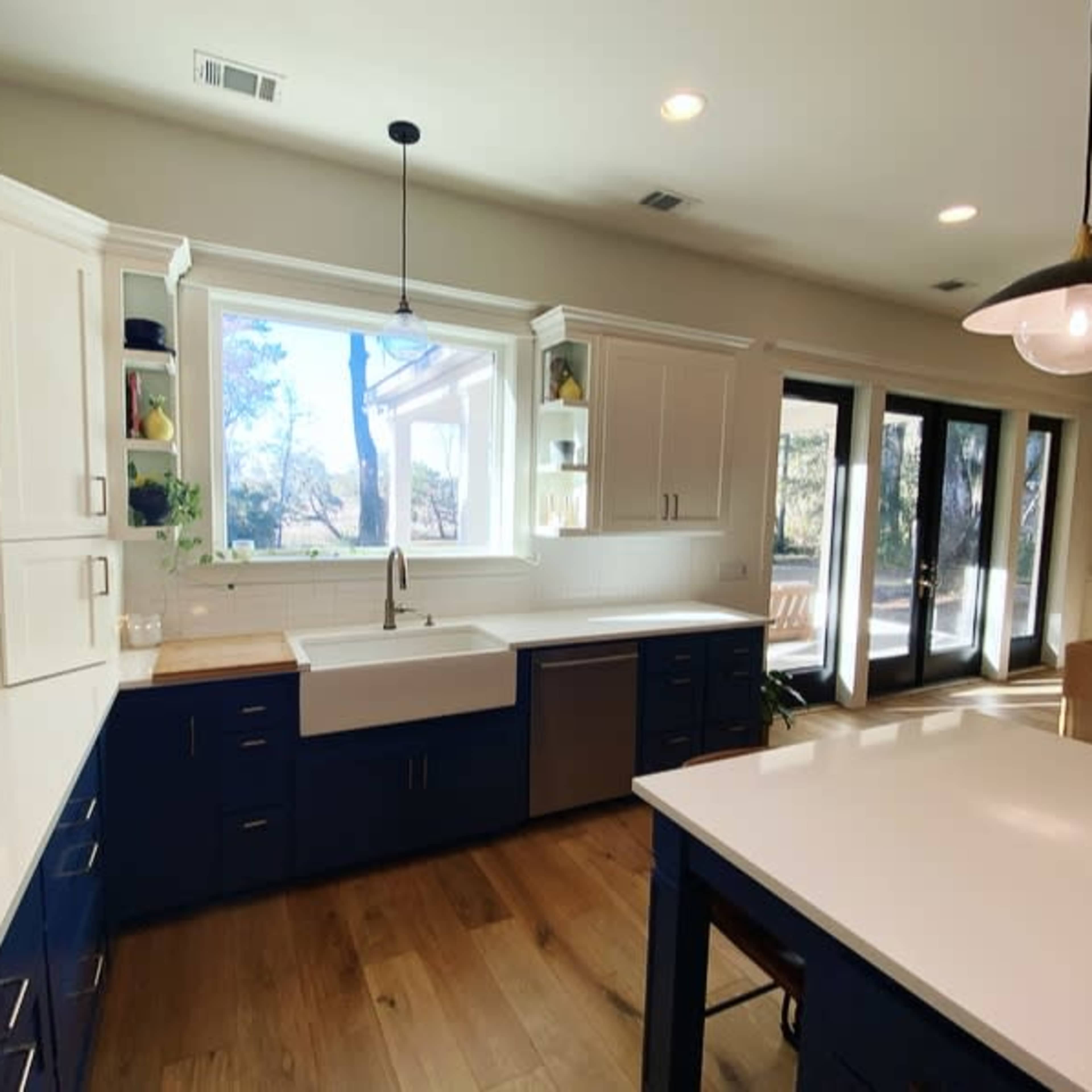 A modern kitchen with white cabinets and blue lower cabinets features a farmhouse sink below a large window and an island with a countertop.