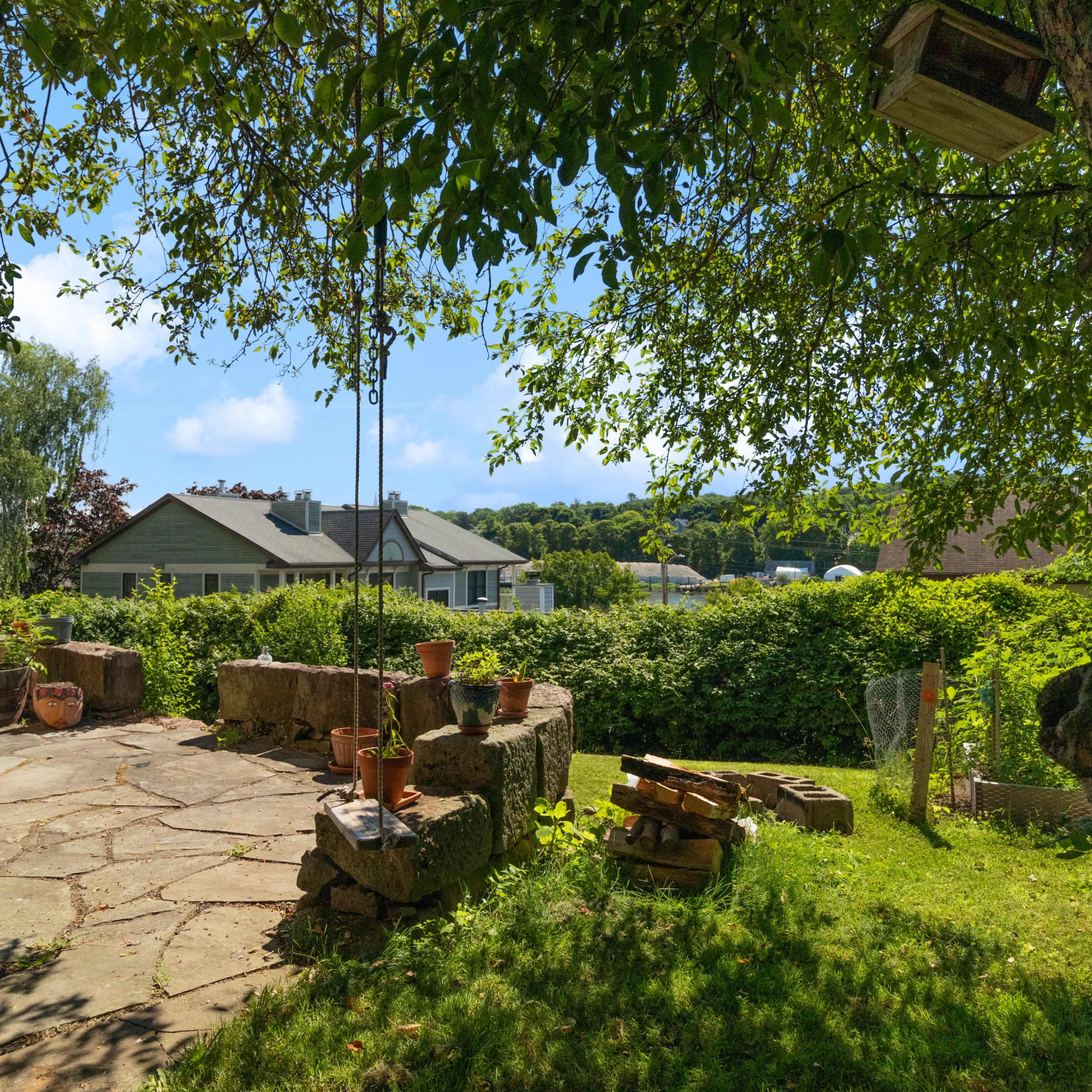 A stone patio surrounded by greenery, with a few potted plants and houses visible in the background under a clear sky.