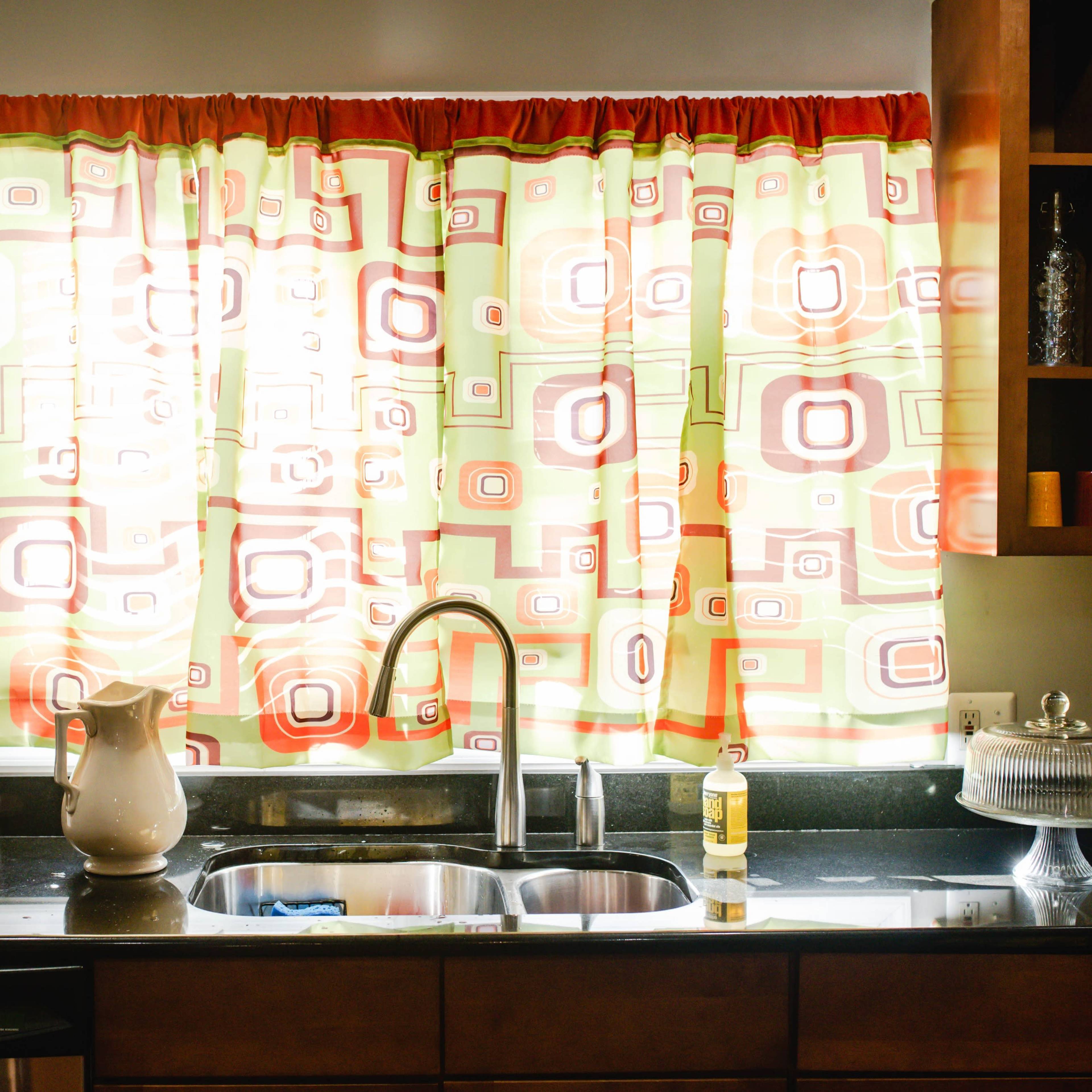 A kitchen features a sink with a dark countertop, illuminated by light filtering through a patterned curtain in shades of green, red, and orange.
