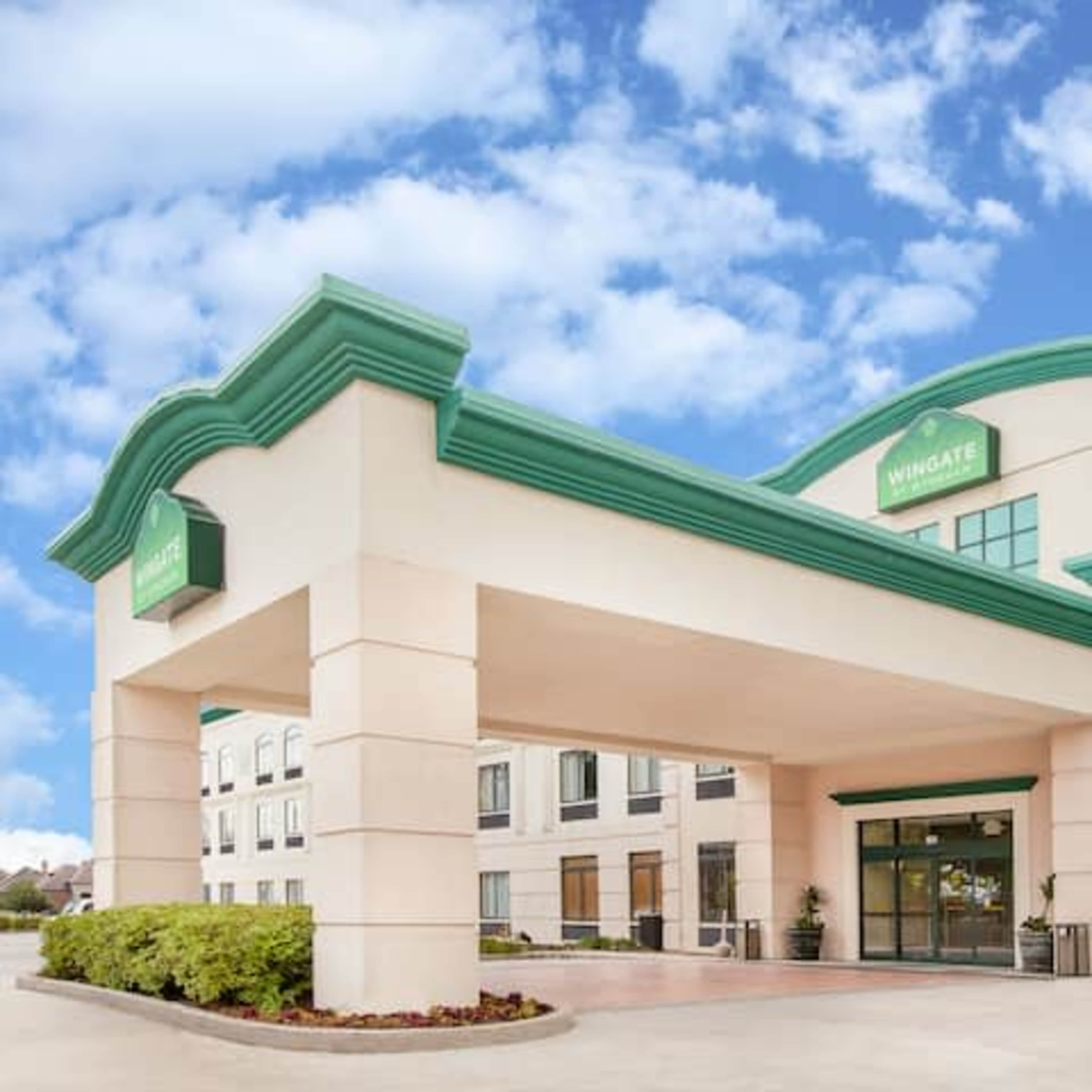 The entrance of a Wingate hotel building with a pale exterior and a green accent roof under a blue sky.