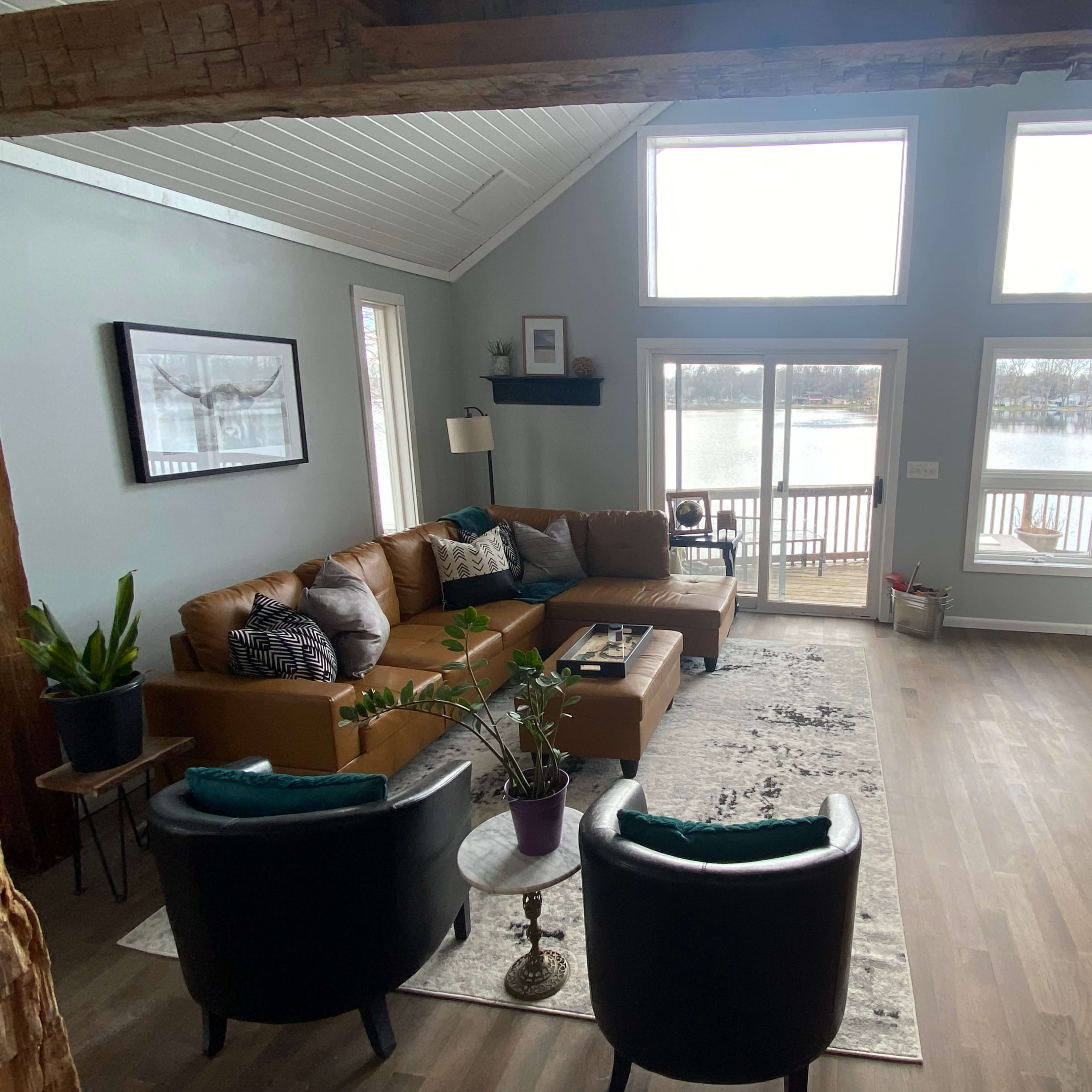 The living room features a brown leather sectional sofa, two black chairs, and large windows that overlook a lake.