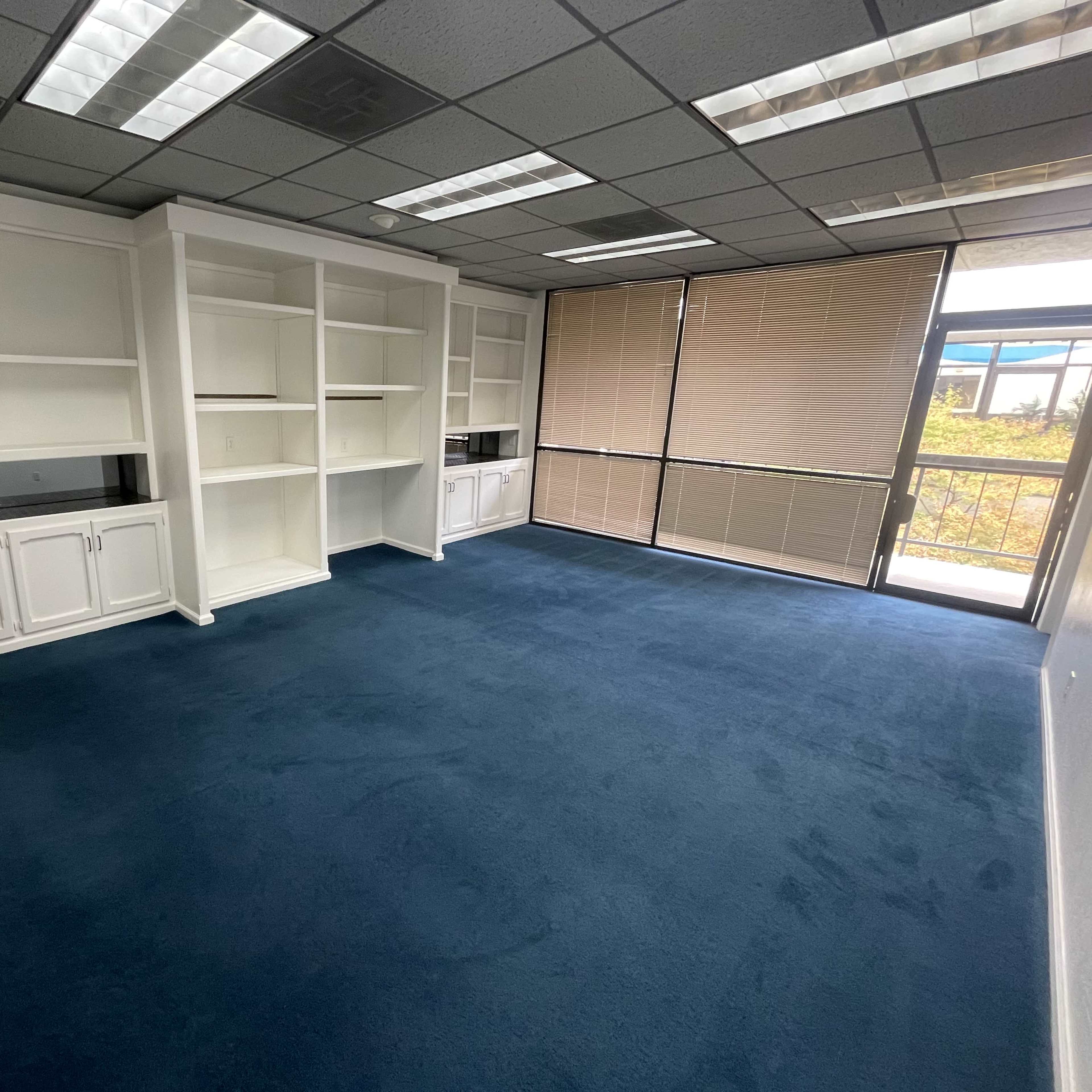An empty office space with blue carpeting, white built-in shelves, and large windows covered by blinds.
