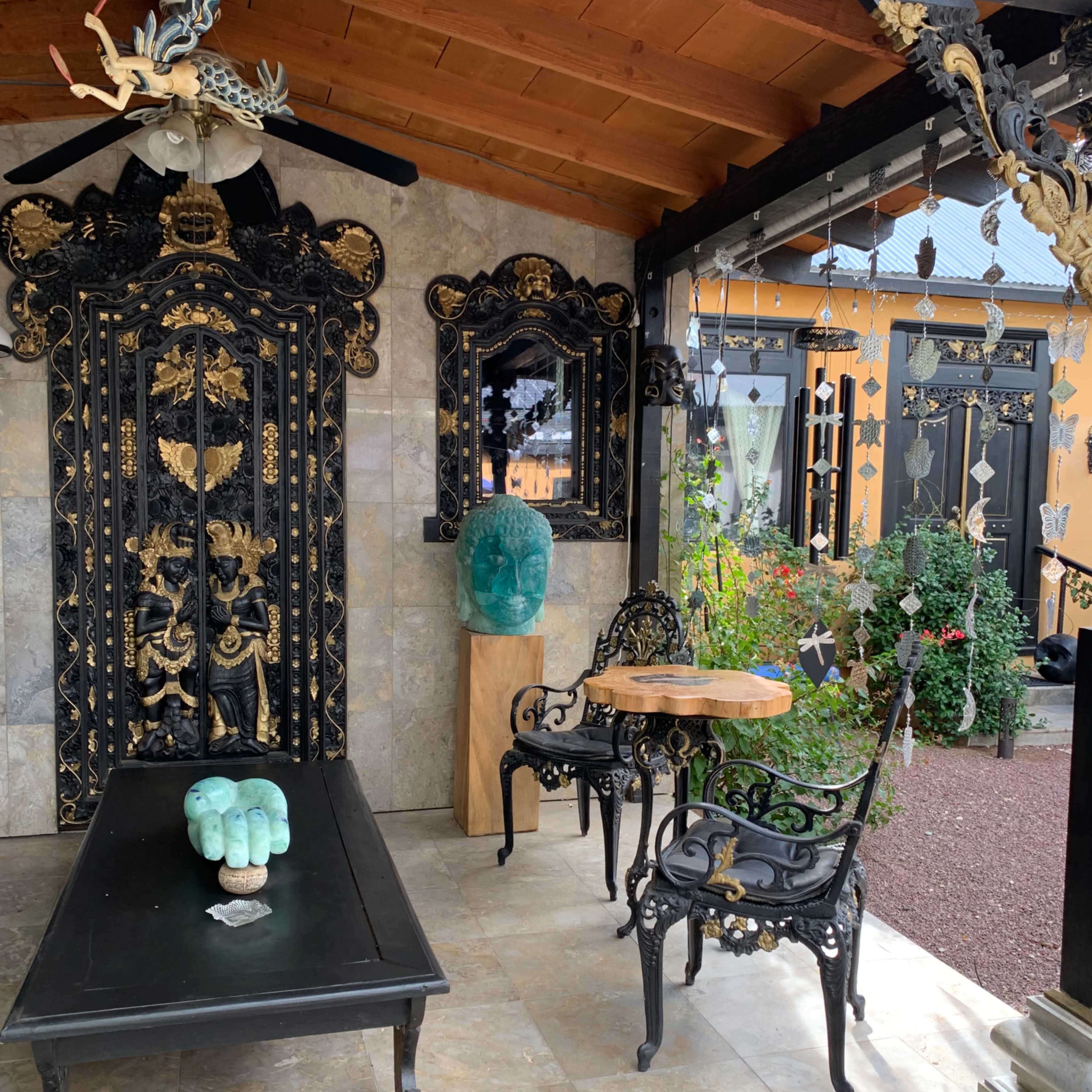 A decorative outdoor space featuring a black intricately carved wooden door, a stone-tiled floor, and wrought iron furniture with a small table and a sculptural green piece on it.