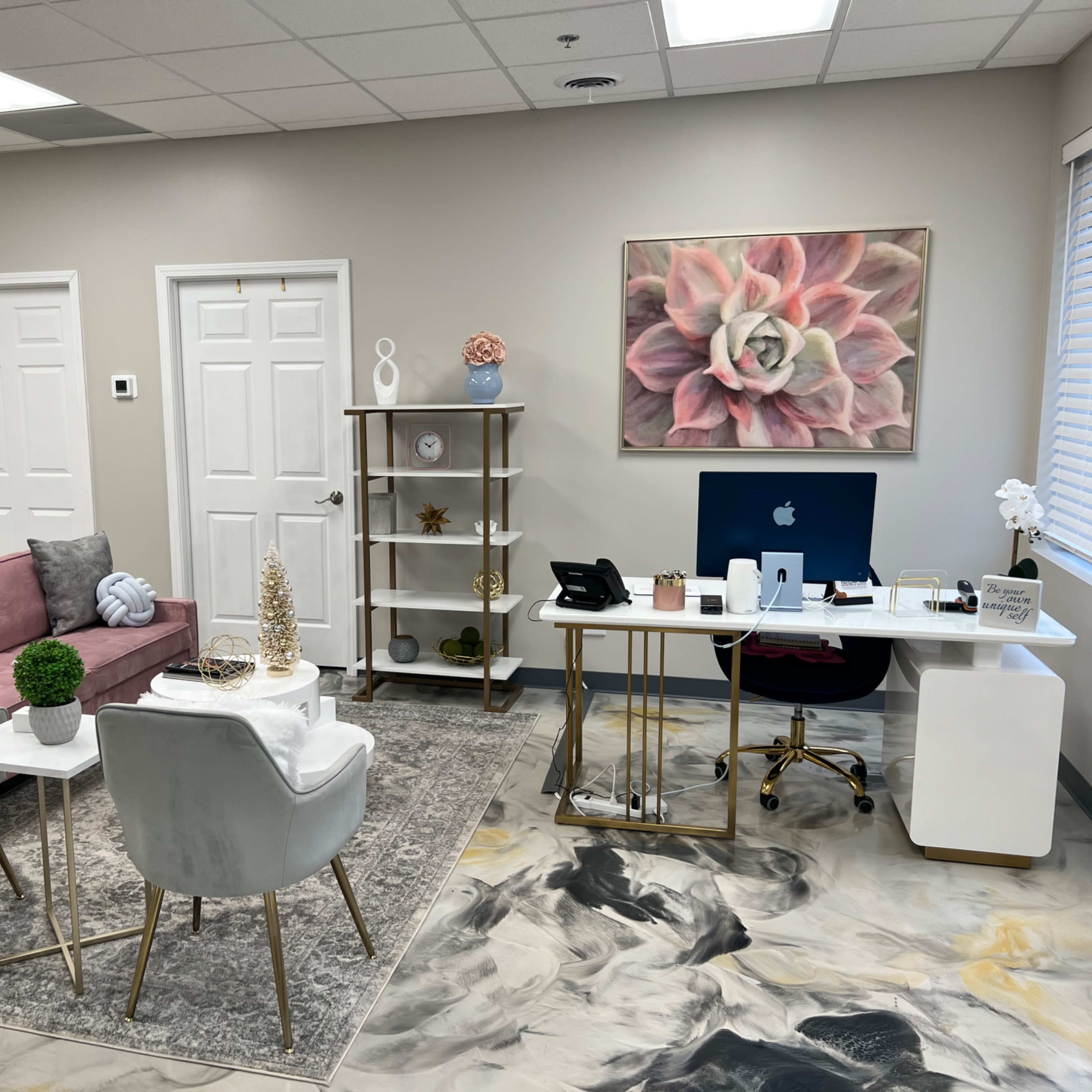 A modern office space featuring a seating area with a pink sofa and two chairs, a decorative bookshelf, and a desk with a computer against a light-colored wall.