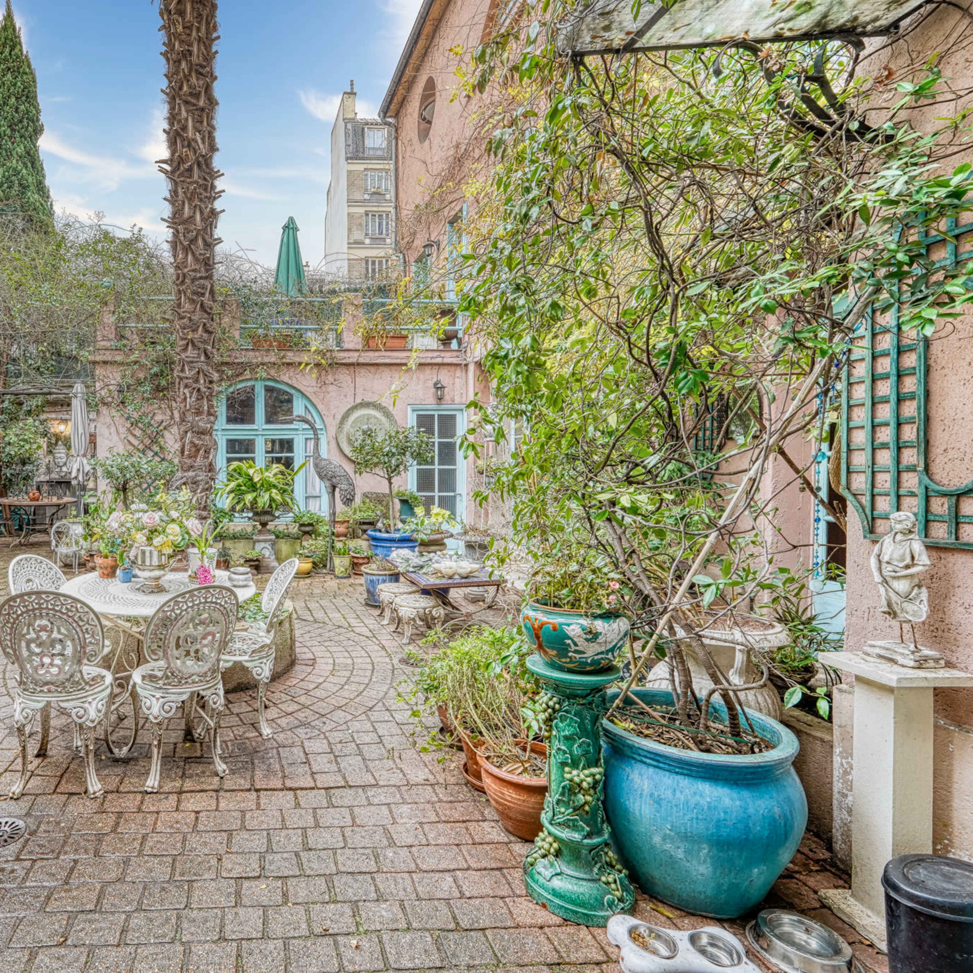 A lush courtyard garden with various potted plants, stone pathways, and a table set with chairs under a palm tree.