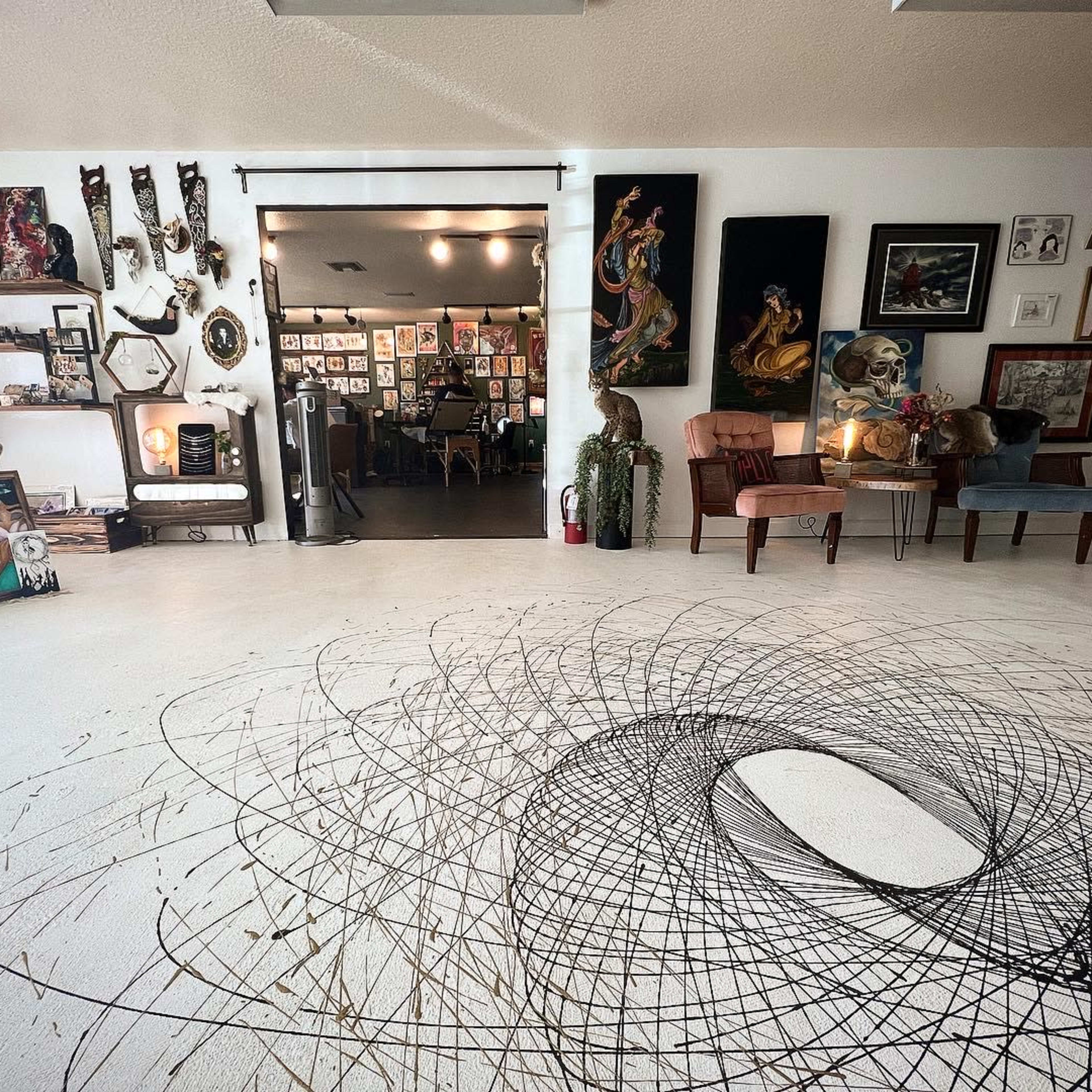 An art studio with a spiraling black pattern on the white floor, surrounded by various framed artworks and decorative items on the walls.