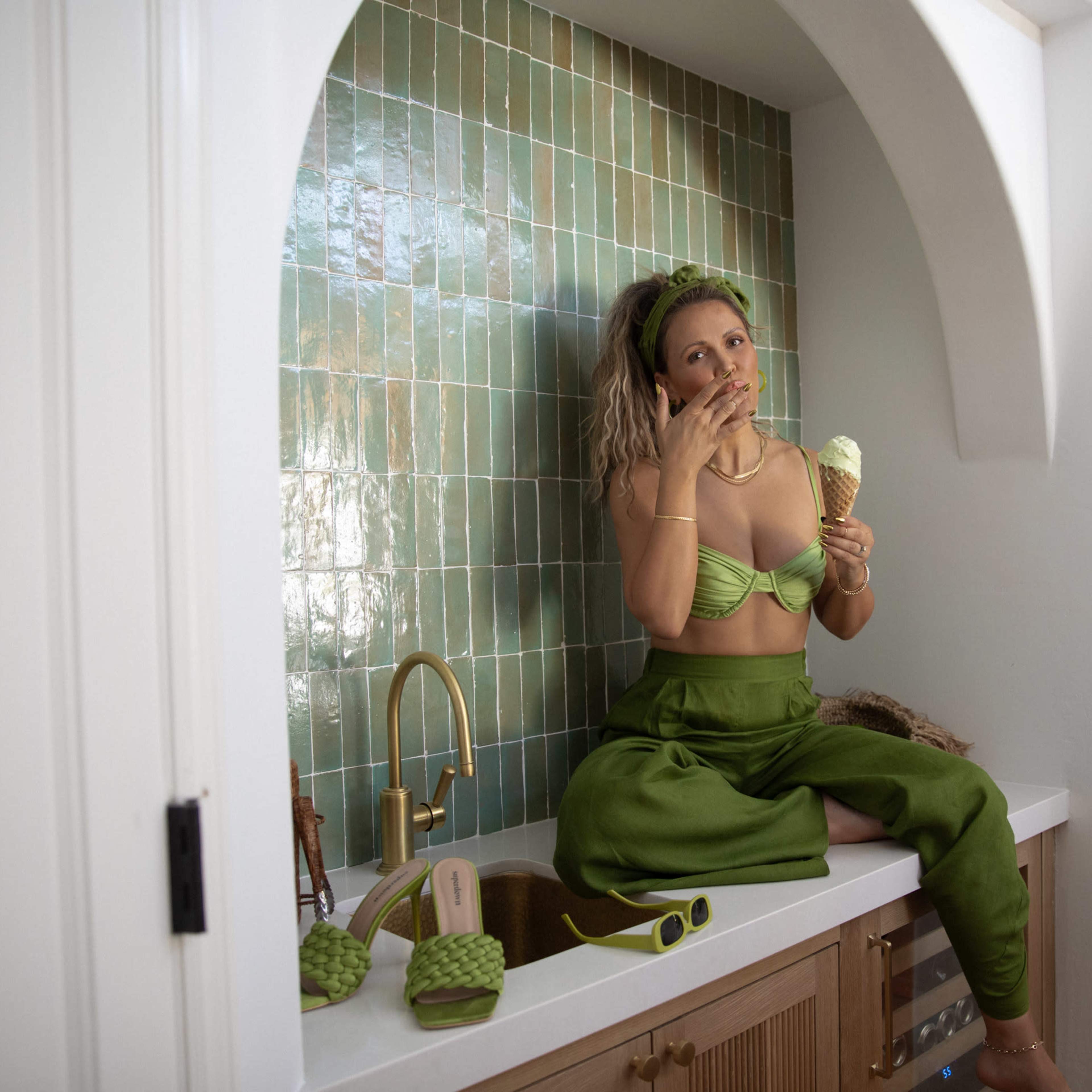 A woman in a green outfit sits on a counter, enjoying an ice cream cone in a brightly lit kitchen with teal tile.