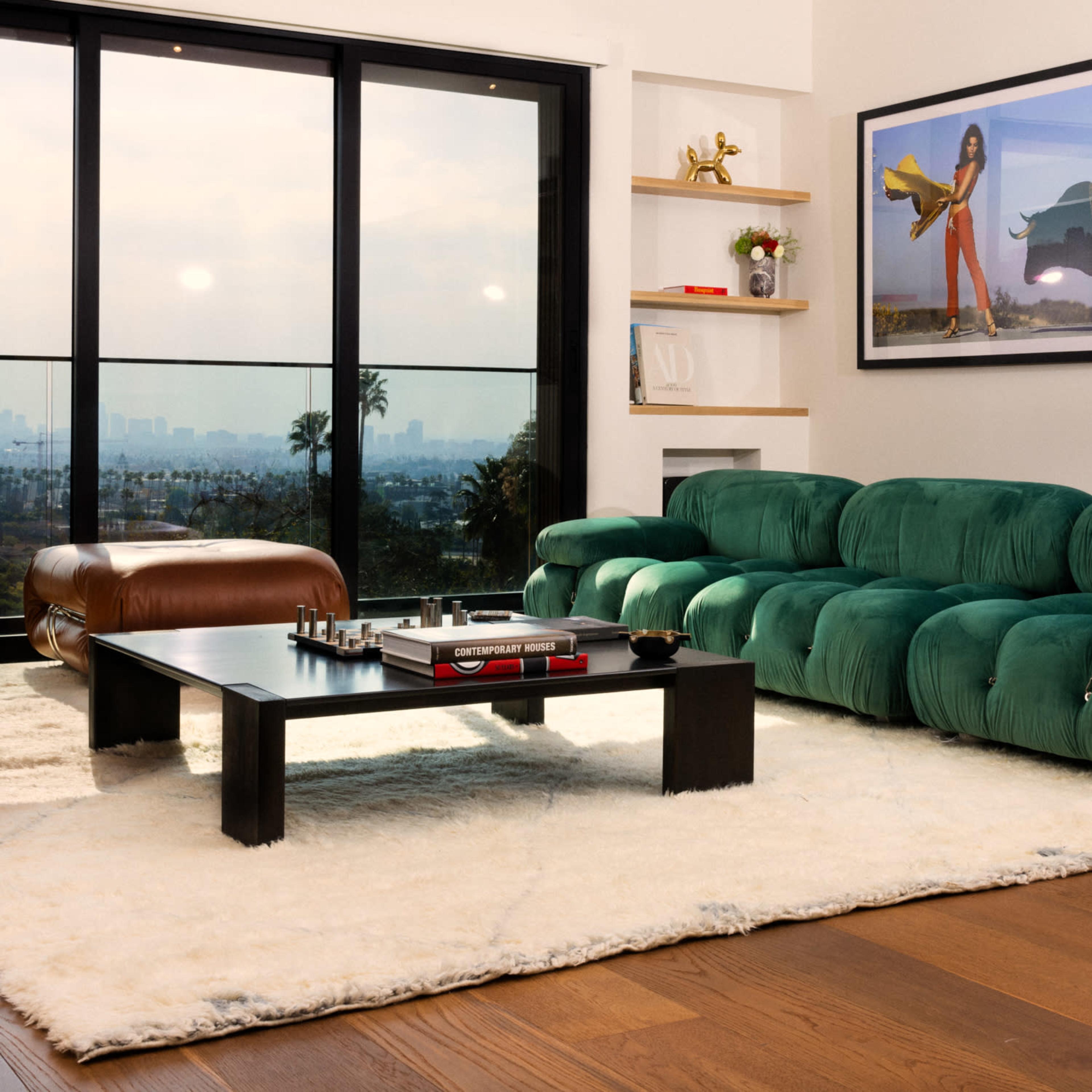 A modern living room with a green sectional couch, a black coffee table, and large windows that overlook a city skyline.
