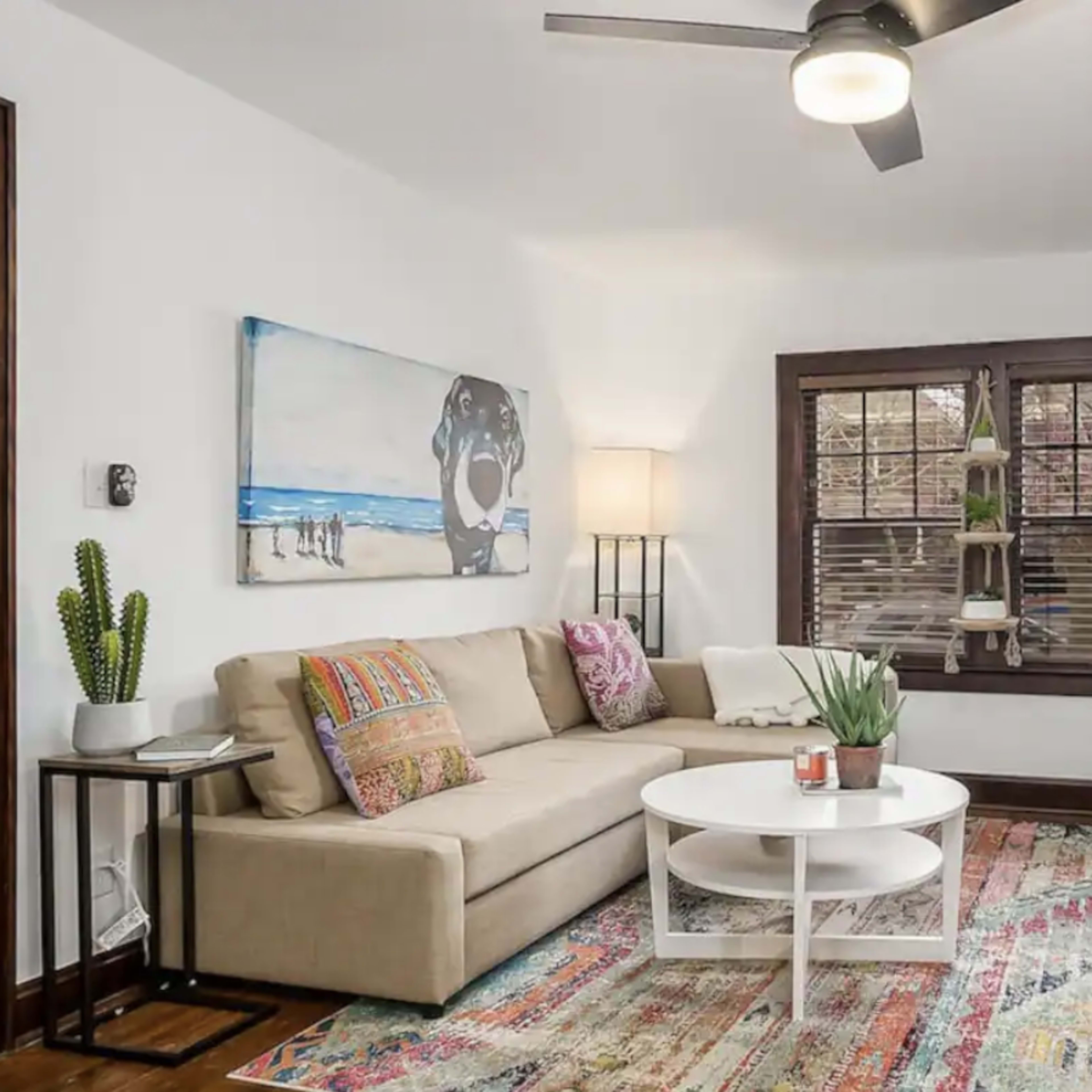 A living room featuring a beige sectional sofa, a round white coffee table, a patterned area rug, and large windows with blinds, complemented by a small indoor plant and decorative items.